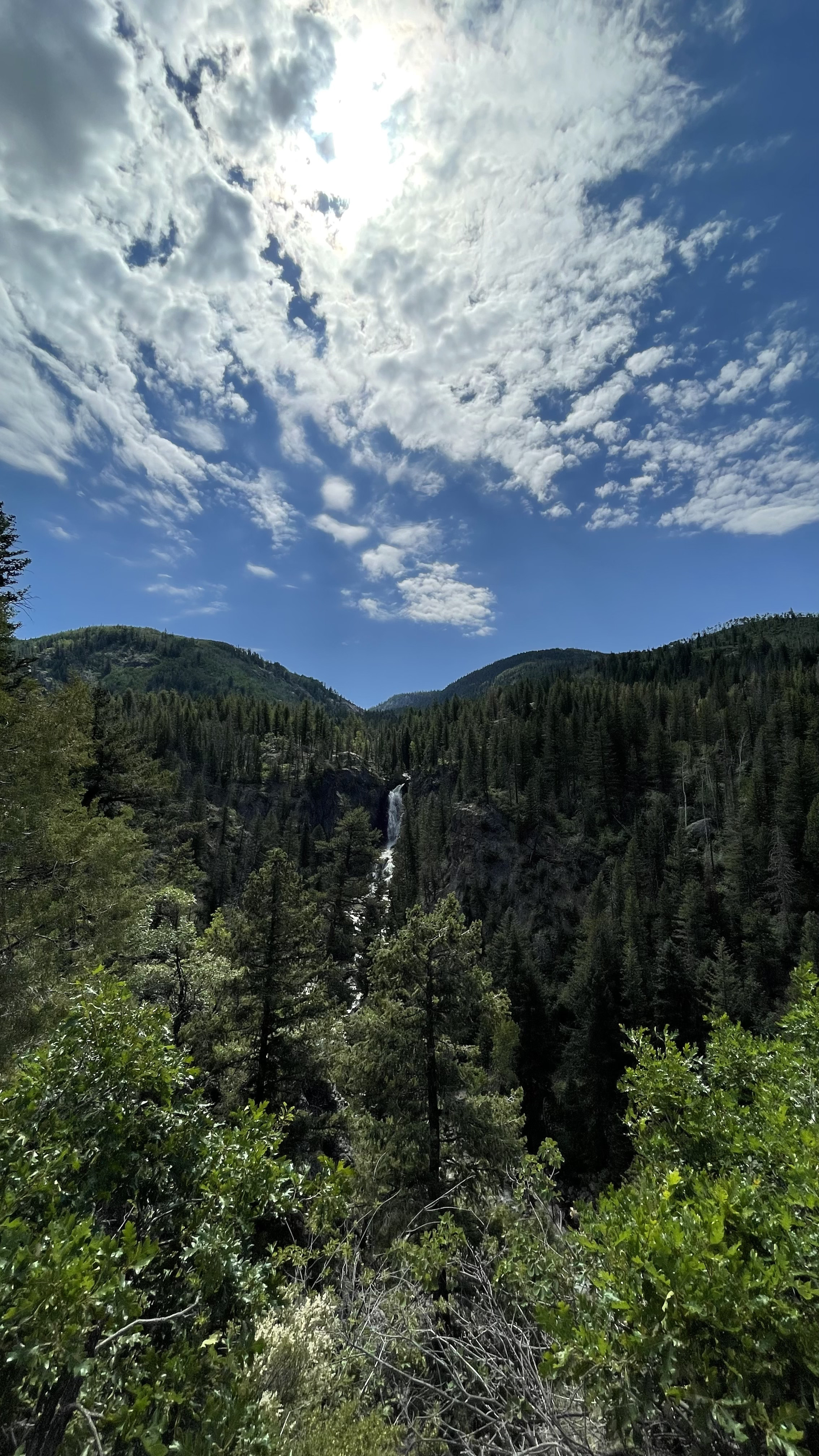 A scenic mountain landscape with a waterfall flowing down a rocky cliff surrounded by dense pine trees, under a partly cloudy sky with the sun shining.