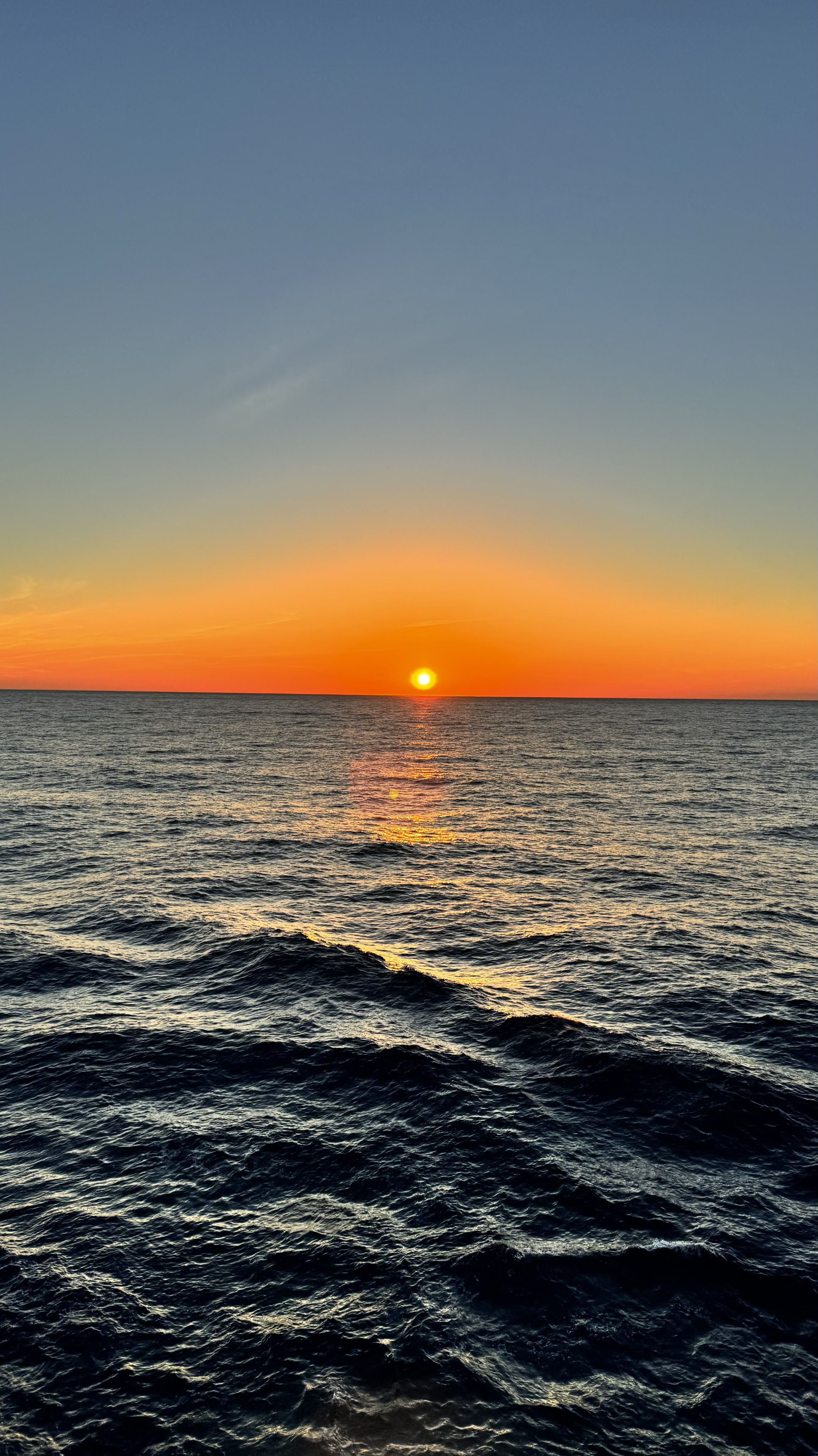 Sunset over the ocean, with the sun near the horizon and calm, wavy water in the foreground.