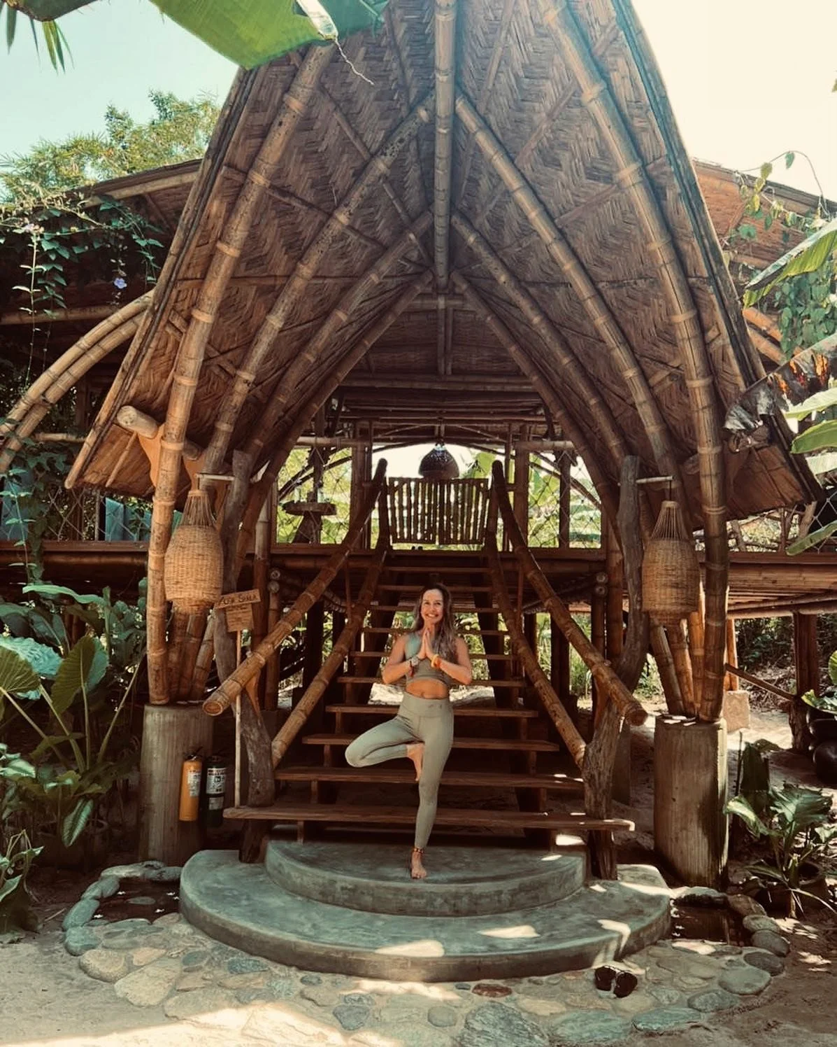 A woman practicing yoga in a tree pose in front of a bamboo hut with stairs, surrounded by tropical plants.