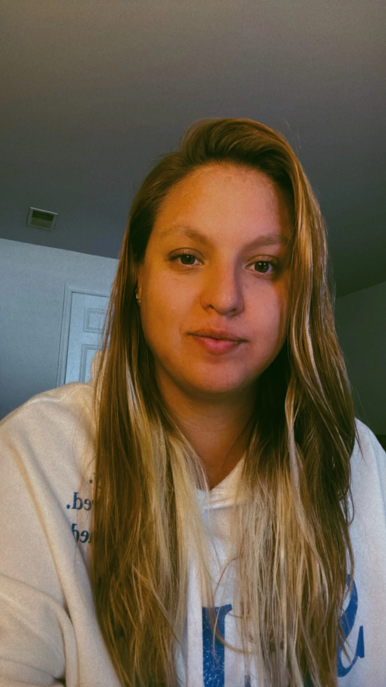 A young woman with long wavy blond hair and light skin, wearing a white hoodie, is looking at the camera. The background shows a closed door and a plain ceiling.