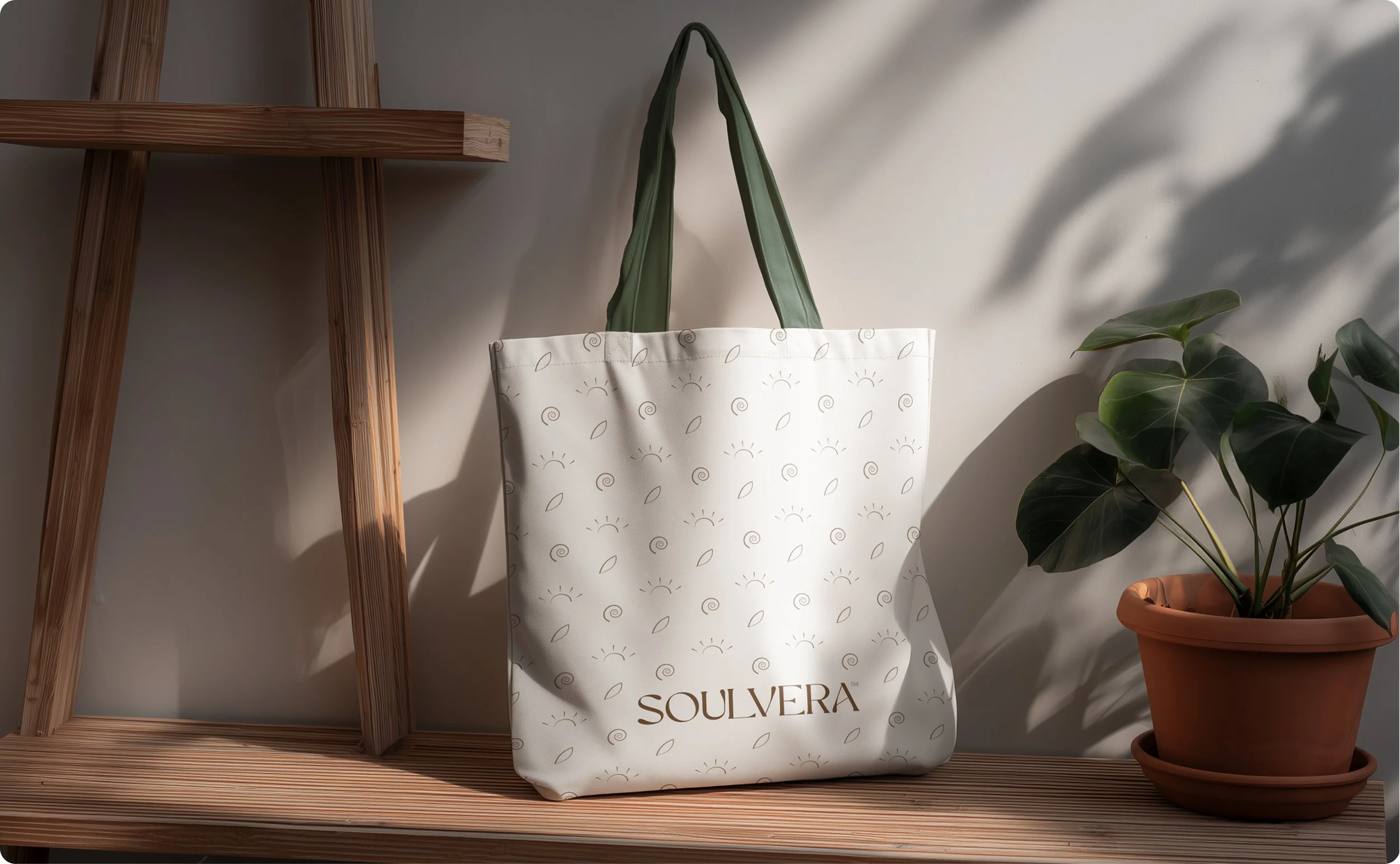 A white tote bag with a sun and leaf pattern, featuring the words 'SOUVERA' and 'OLIVE' printed on it, is placed on a wooden surface with a wooden ladder on the left and a potted olive plant on the right.