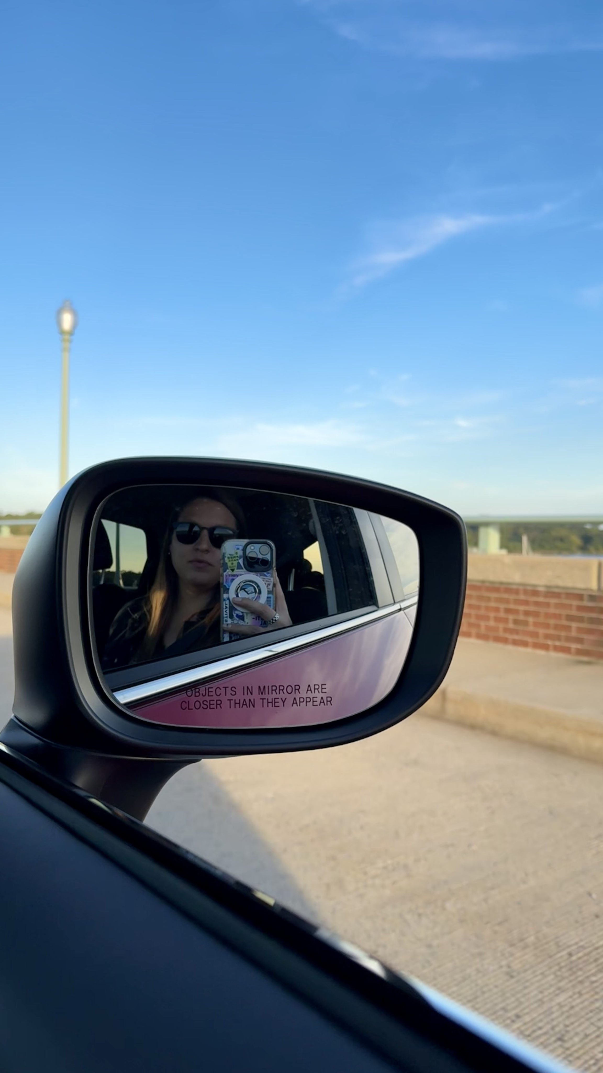 A woman wearing sunglasses is taking a selfie with her phone, which has a colorful case, in the side mirror of a car.