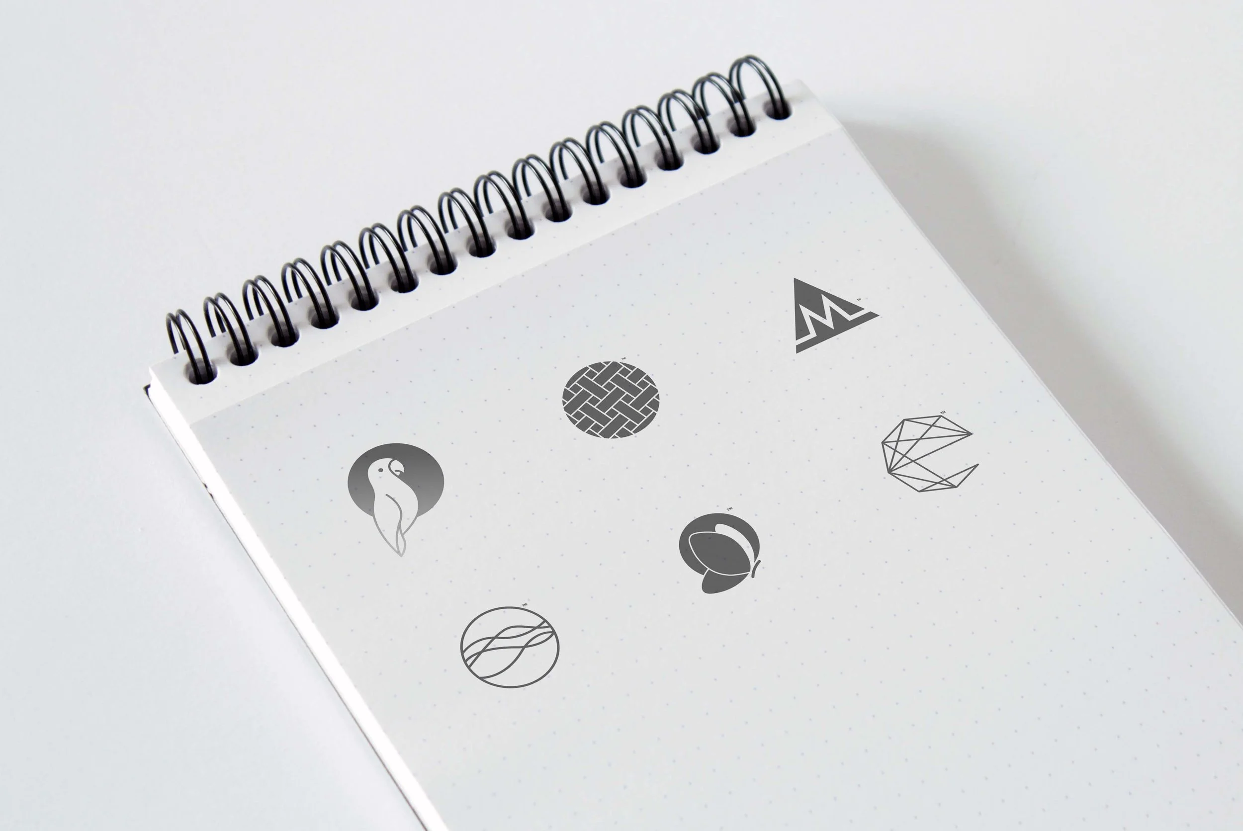 Close-up of a white spiral-bound notepad with black rings, featuring various black and white icons including a maze, a mountain, a penguin, a diamond, and a stylized leaf, on a white background.