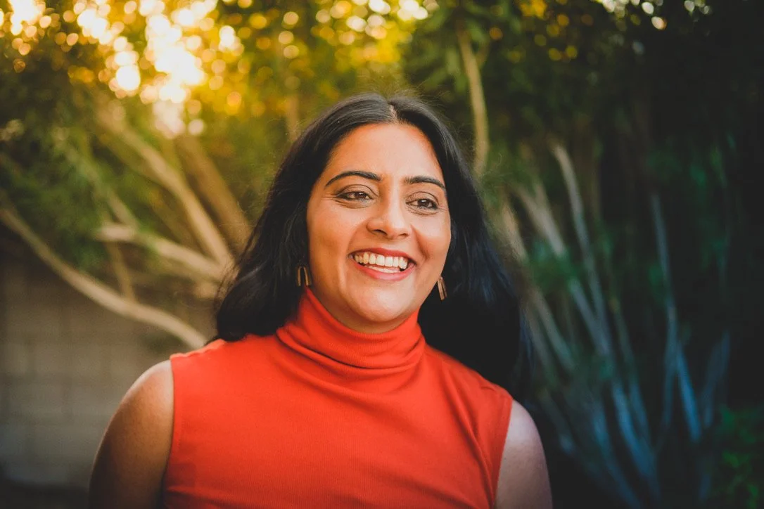 Smiling woman with black hair wearing a red sleeveless top outdoors with trees and sunlight in the background.