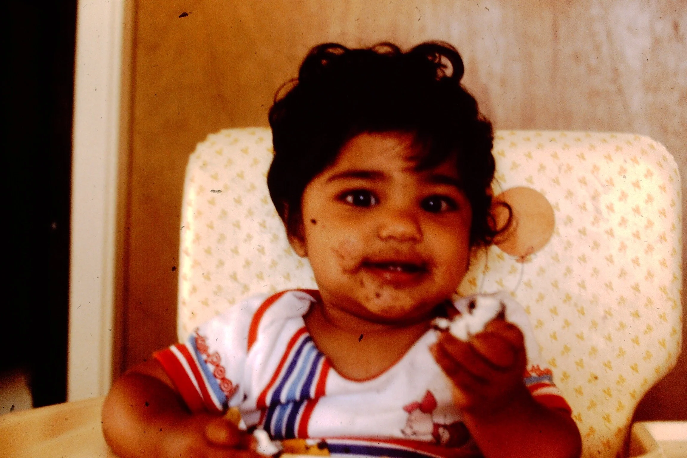 A young child with curly dark hair and baggy cheeks is sitting in a high chair, holding and eating a piece of food. The child is smiling and has some food or chocolate smudges on their face. The high chair has a pattern with small yellow and pink designs, and the background appears to be a beige wall.