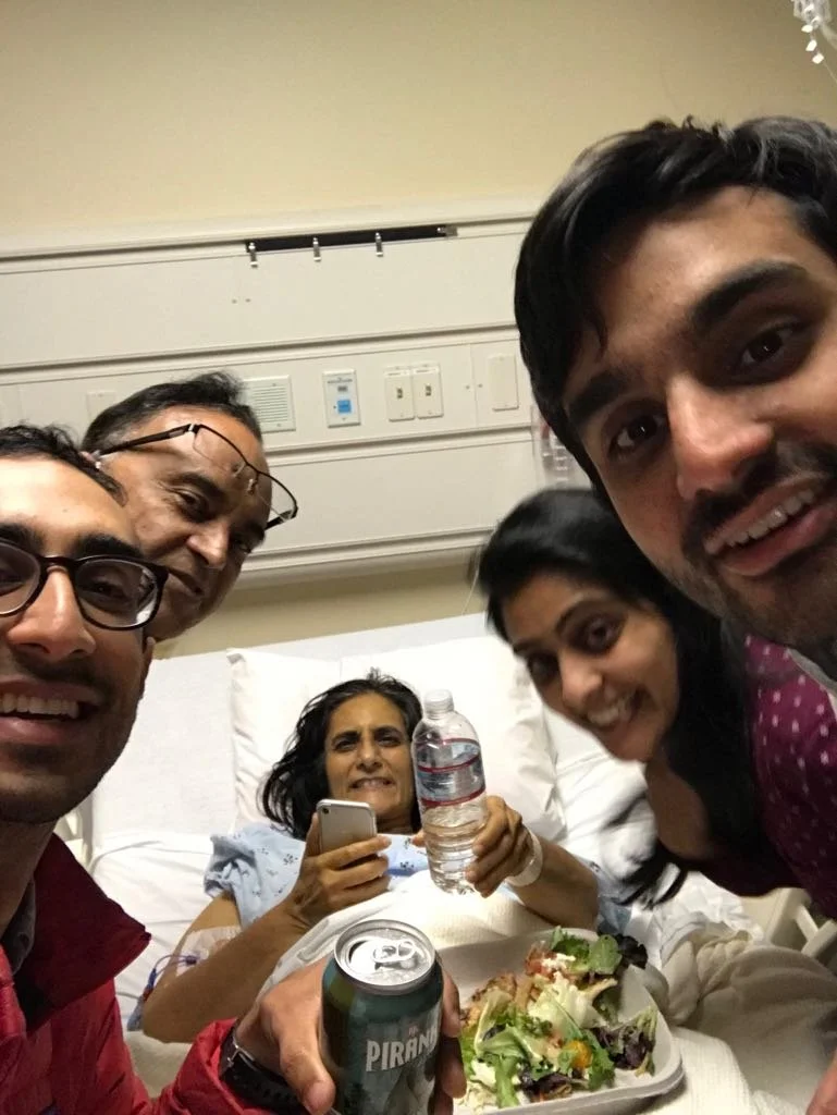 Group of five people taking a selfie in a hospital room, with a woman in a hospital bed holding a water bottle and phone, surrounded by friends holding a soda can, water bottle, and salad, smiling.