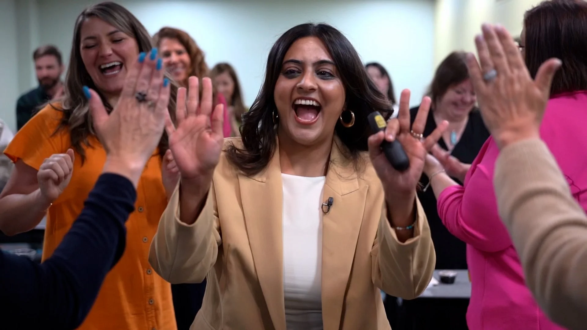 A woman with dark hair, wearing a beige blazer and white top, smiling and raising her hands in a high-five with people around her in a lively group setting.