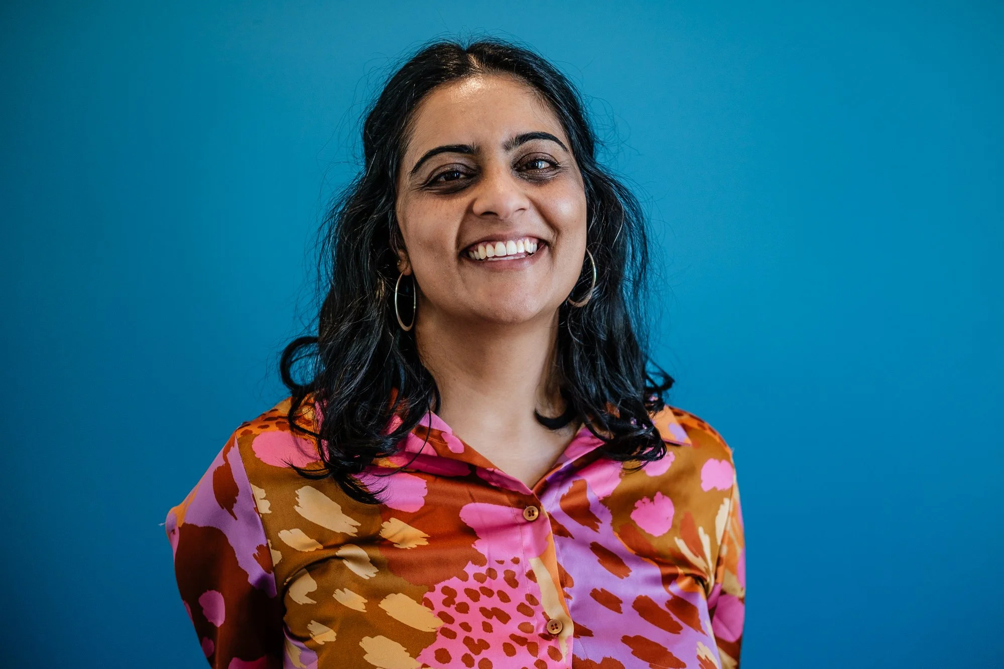 A smiling woman with dark, wavy hair in front of a blue background, wearing a colorful patterned shirt with pink, orange, and beige tones, and hoop earrings.