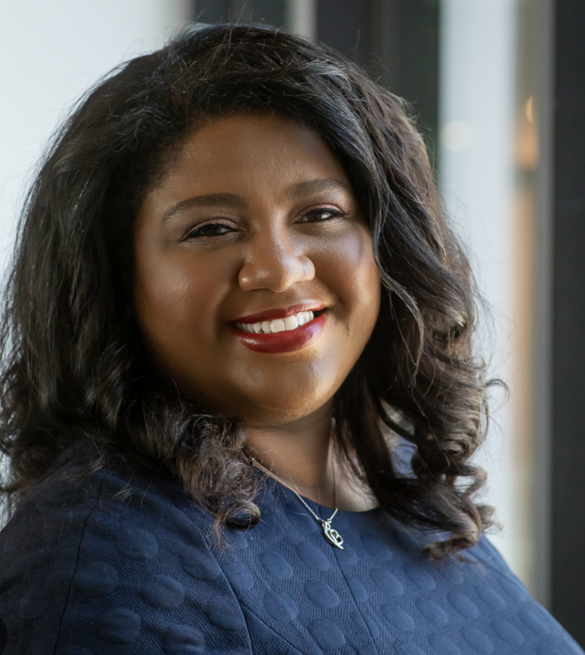 A smiling woman with dark curly hair, wearing a navy blue dress and a silver necklace, standing indoors near a window.
