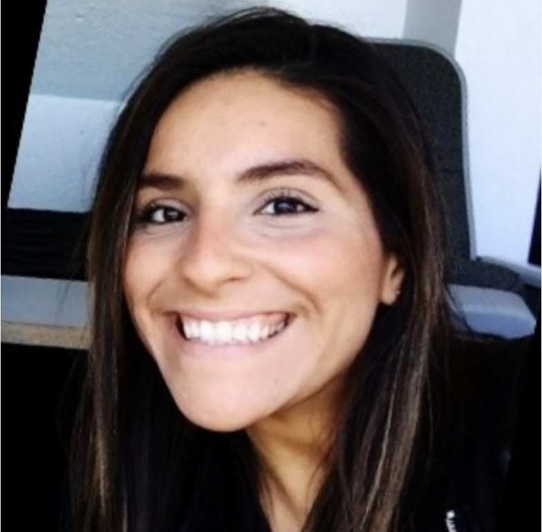 A young woman with long dark hair smiling at the camera, sitting indoors.