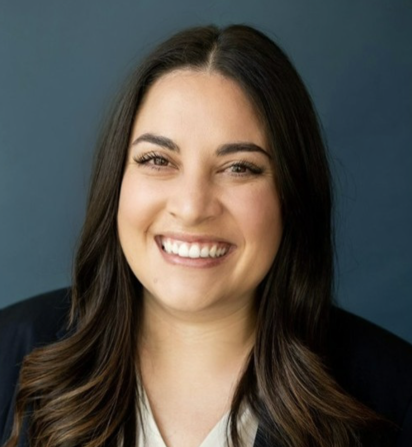 Headshot of a smiling woman with long dark hair, wearing a dark blazer, against a blue background.