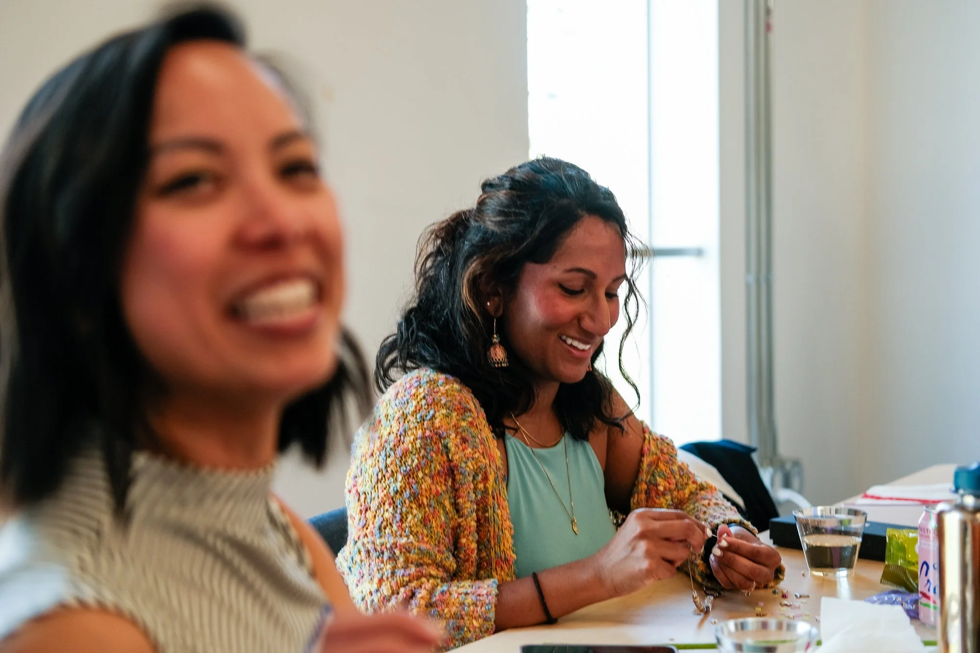 Two women sitting at a table, smiling and working on beaded jewelry. One woman is in the foreground, slightly blurred, and the other woman is in the background, focused on her craft.