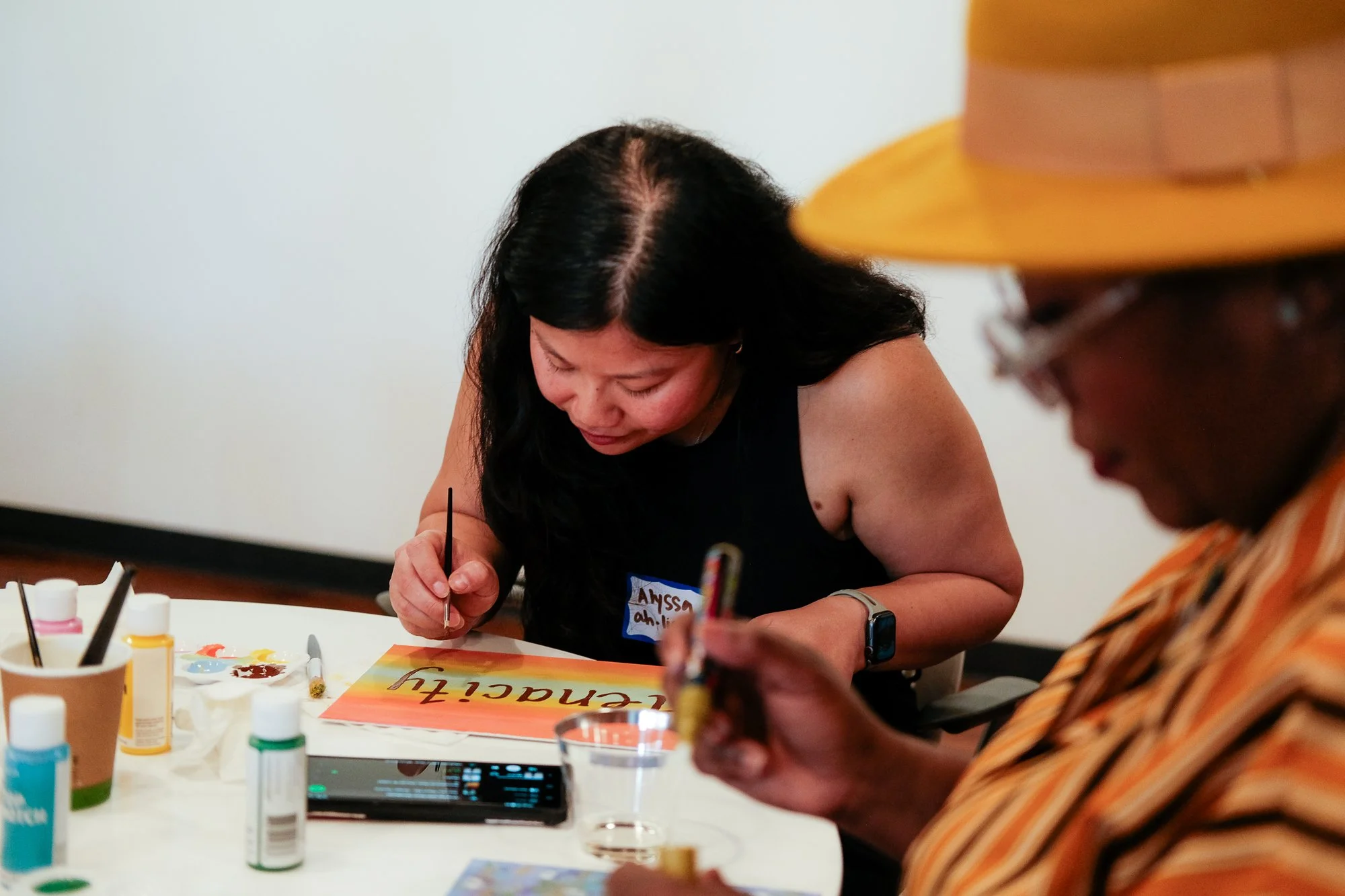 Two people sitting at a table engaged in painting or calligraphy. The woman in the center has black hair and is wearing a black sleeveless top, focusing on her artwork. The man on the right, wearing glasses and a wide-brimmed hat, is also working on art. The table holds various art supplies, including paints, brushes, and a water cup.