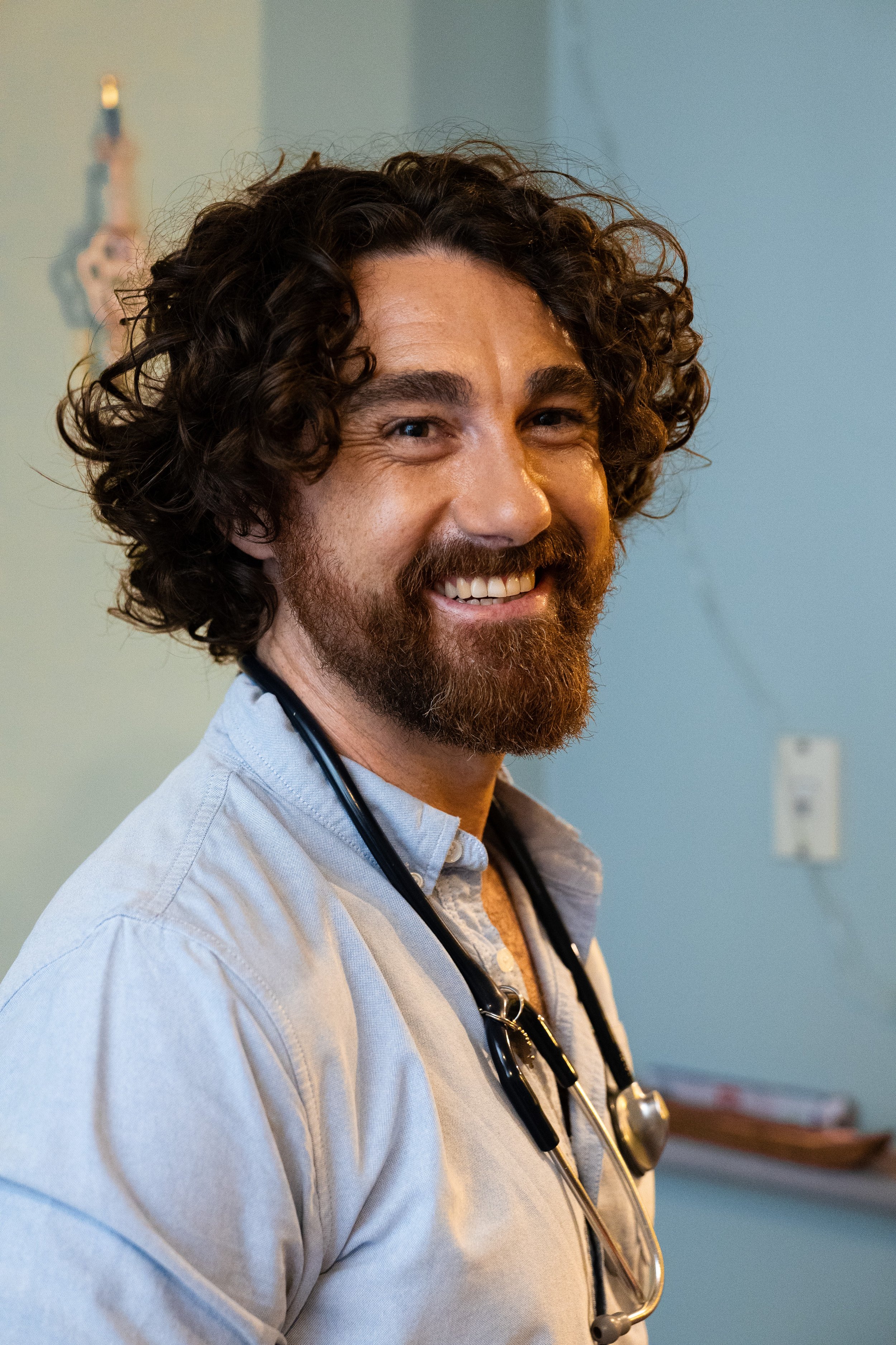 A smiling male doctor with curly brown hair and a beard, wearing a light blue shirt and a stethoscope around his neck, in a medical office or hospital setting.
