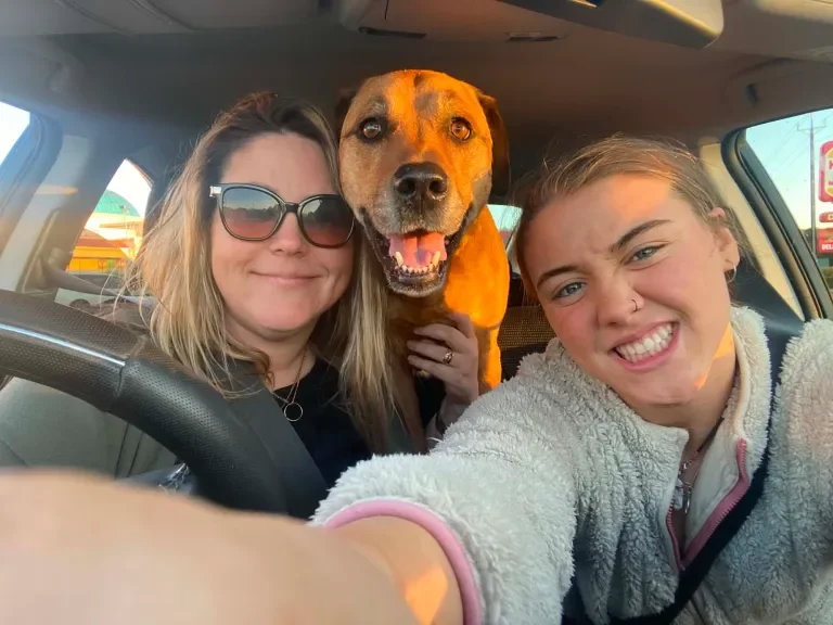 Two women and a dog inside a car, with the women taking a selfie. The woman on the left wears sunglasses and the woman on the right has her hair tied back and is smiling.
