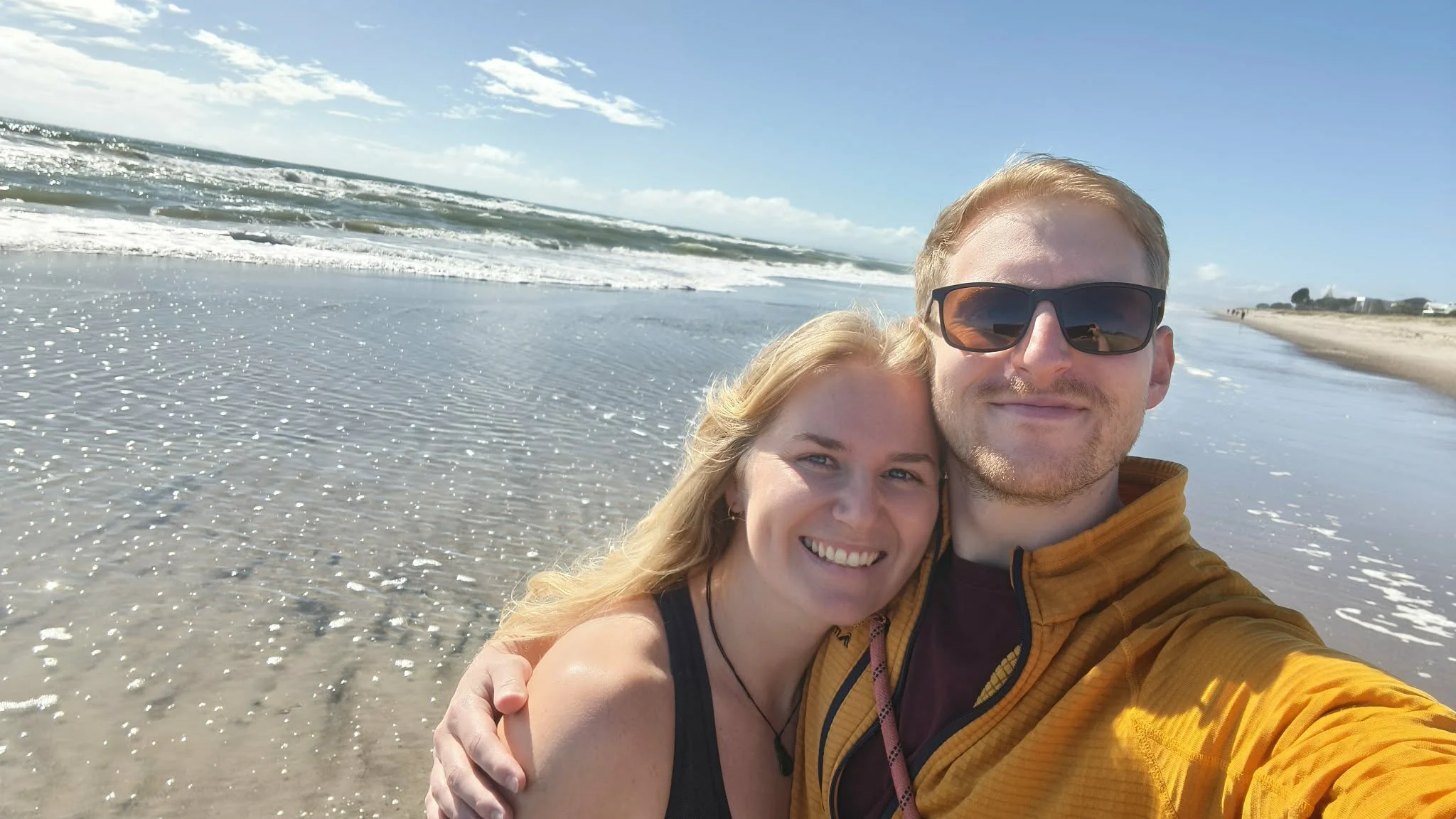 A smiling couple takes a selfie on the beach with waves and blue sky in the background.
