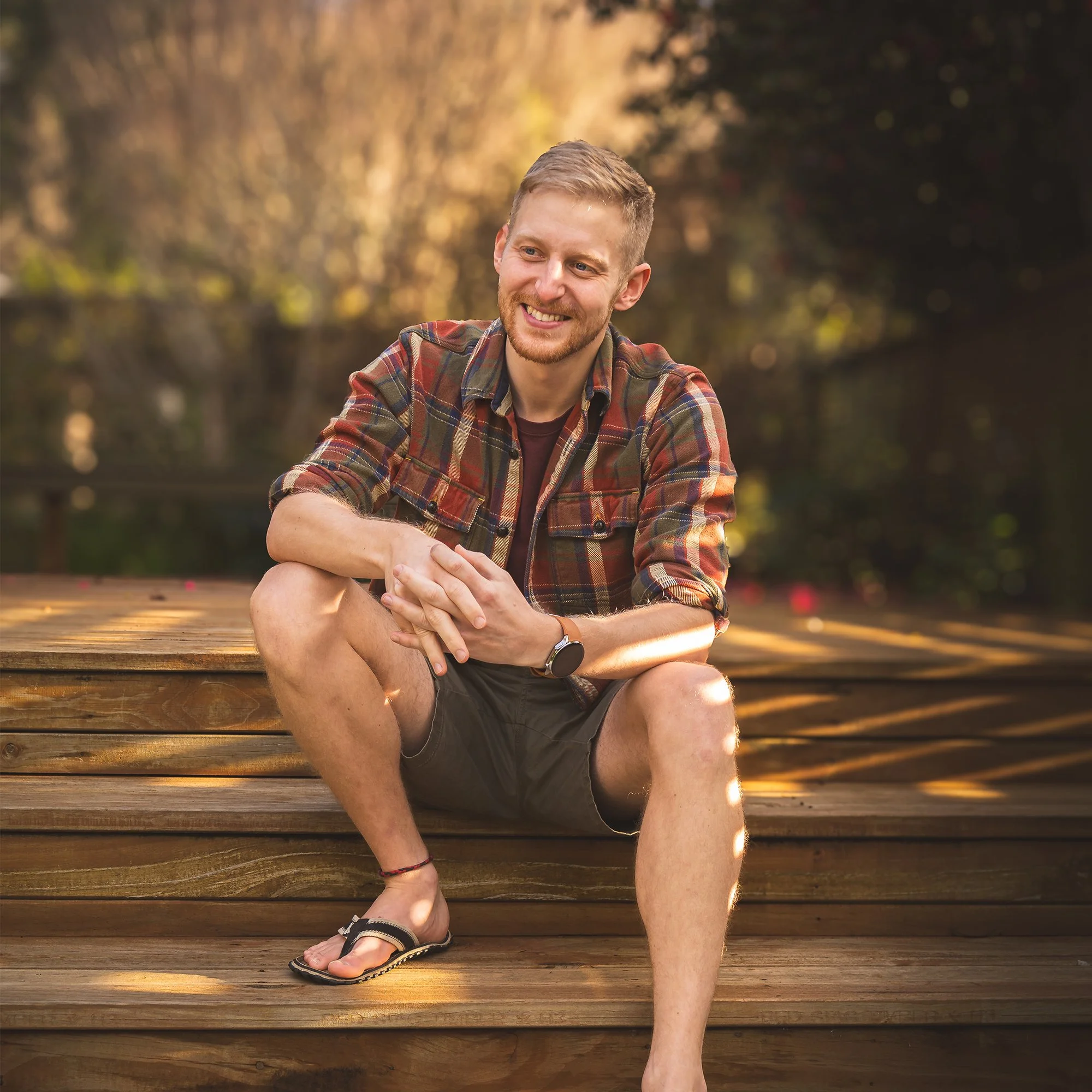A young man with short blond hair and a beard, smiling while sitting on wooden steps outdoors during the daytime. He is wearing a plaid shirt, black shorts, and sandals, with a smartwatch on his left wrist. The background shows a natural setting with trees and a soft, warm glow from sunlight.