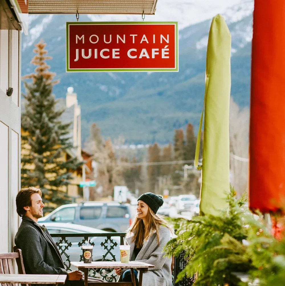 A man and a woman sitting at a table outside Mountain Juice Café, enjoying drinks and smiling at each other on a cloudy day with mountains and trees in the background.