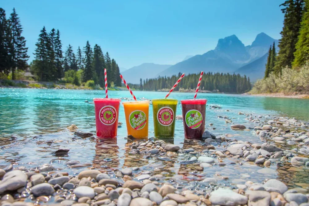Four colourful smoothies from Mountain Juice Cafe in Canmore, with striped straws in a river with pebbles, surrounded by trees and mountains in the background.