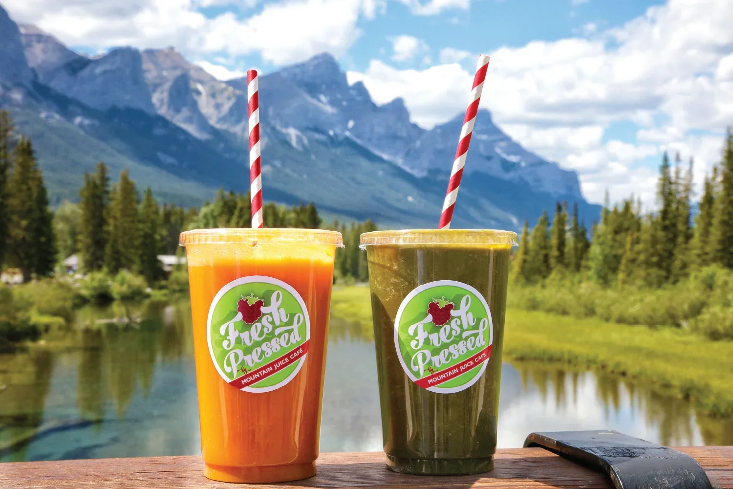 Two cups of Mountain Juice Cafe juice with striped straws sitting on a wooden surface, with a mountain and forest landscape in the background.