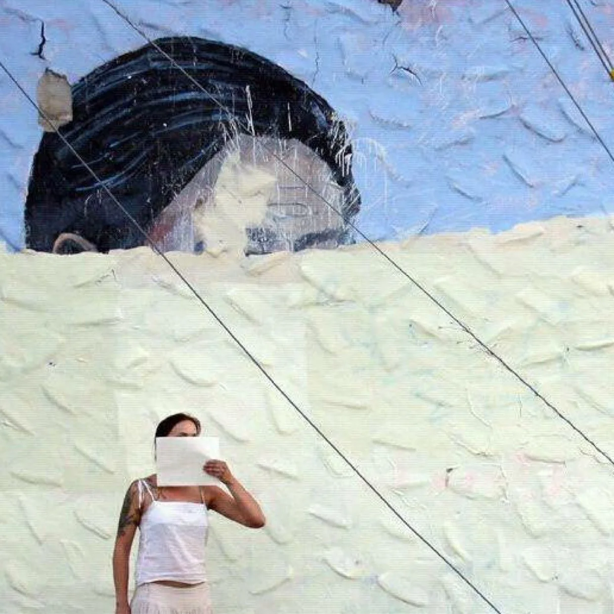 A person holding a sheet of paper in front of her face while standing in front of a large mural of a woman's face that has been partially painted over.