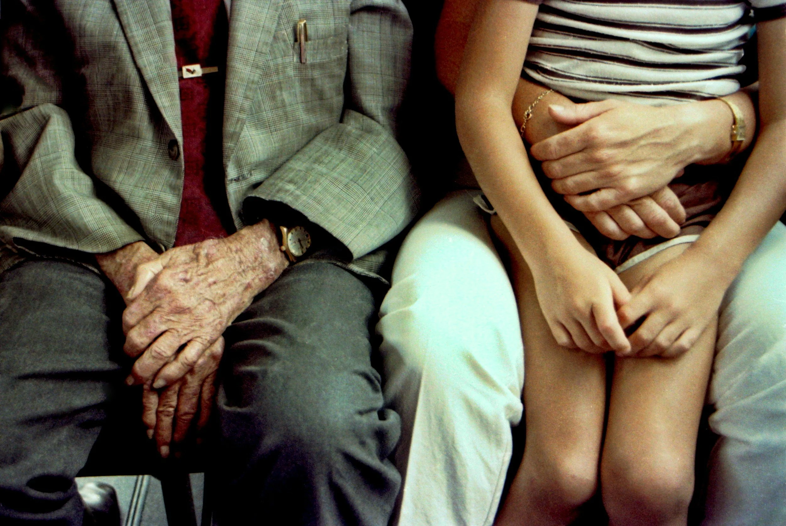Close-up of elderly hands clasped together resting on lap, with middle-aged hands gently holding them, showing a moment of connection and support.