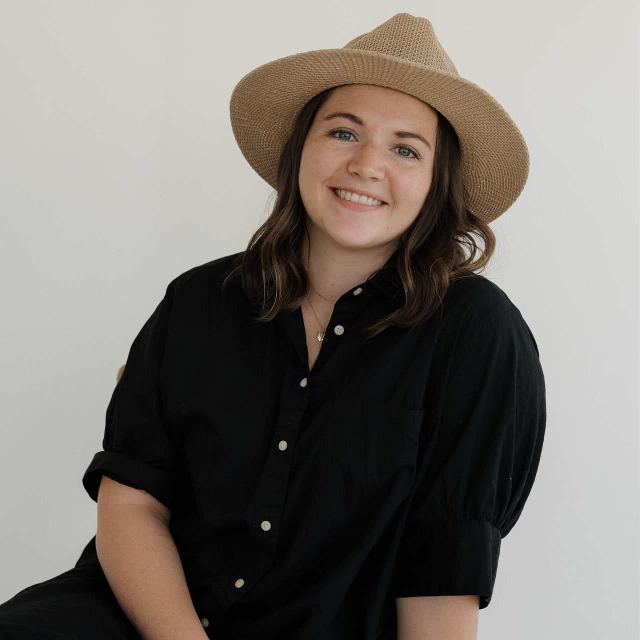 A woman with shoulder-length wavy brown hair wearing a beige straw hat and a black button-up shirt, smiling against a plain white background.
