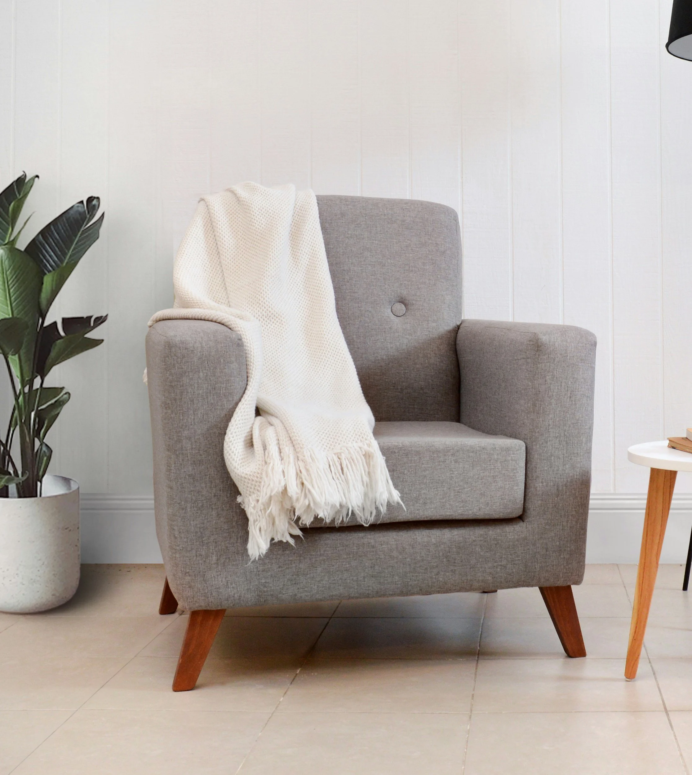 A mid-century modern armchair with a gray fabric upholstery and wooden legs, with a cream-colored chunky knit throw blanket draped over one arm, placed next to a large potted plant and a small white side table with books, against a white paneled wall.