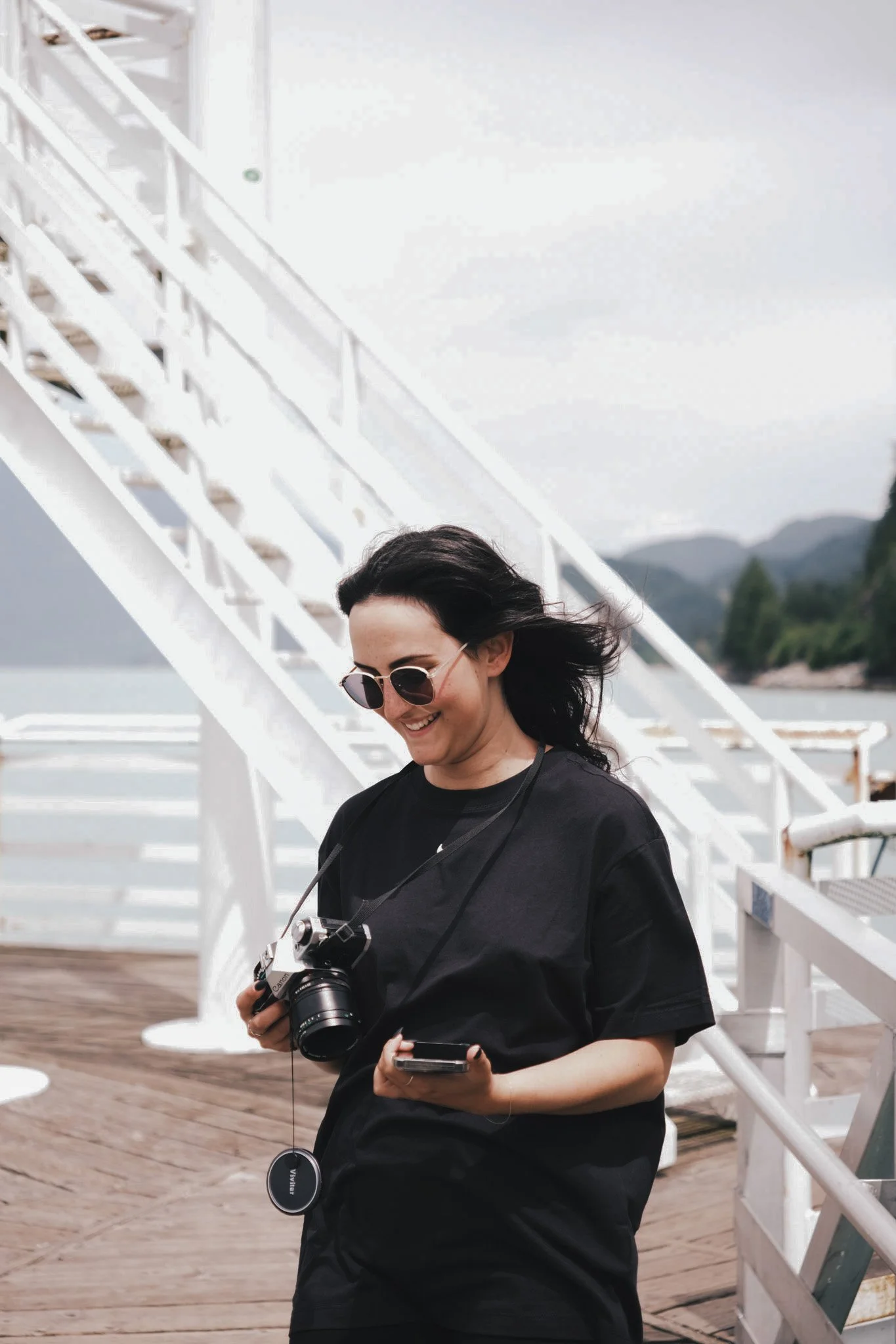 A woman with dark hair, wearing sunglasses and a black shirt, smiling while holding a camera and a smartphone, standing outdoors on a wooden deck near a white staircase with mountains in the background.