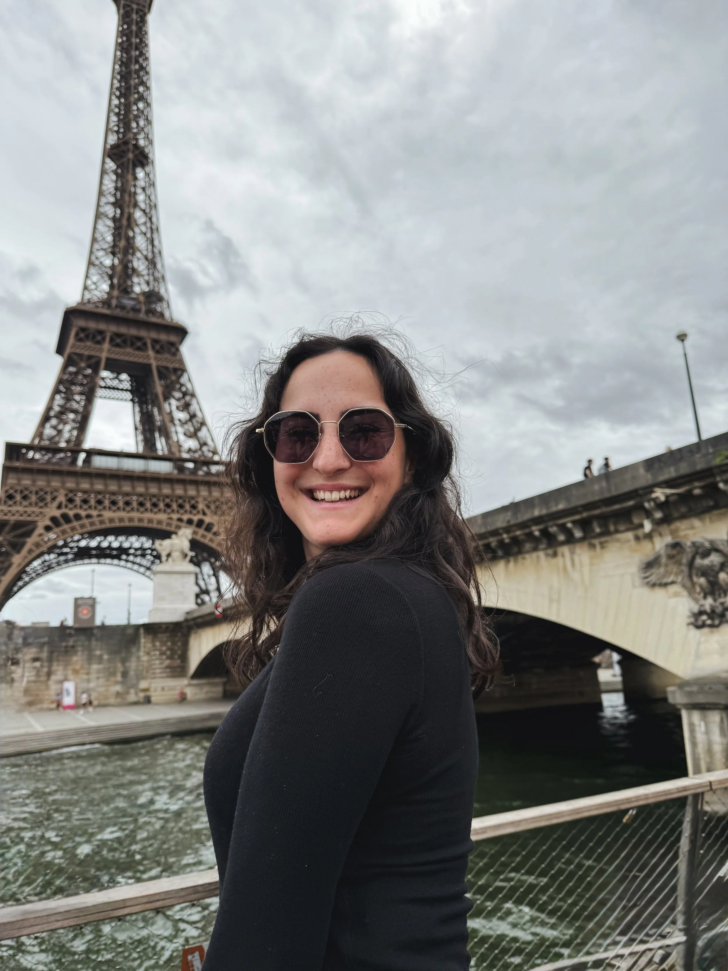 A woman smiling in front of the Eiffel Tower and the Seine River in Paris, France, during overcast weather.