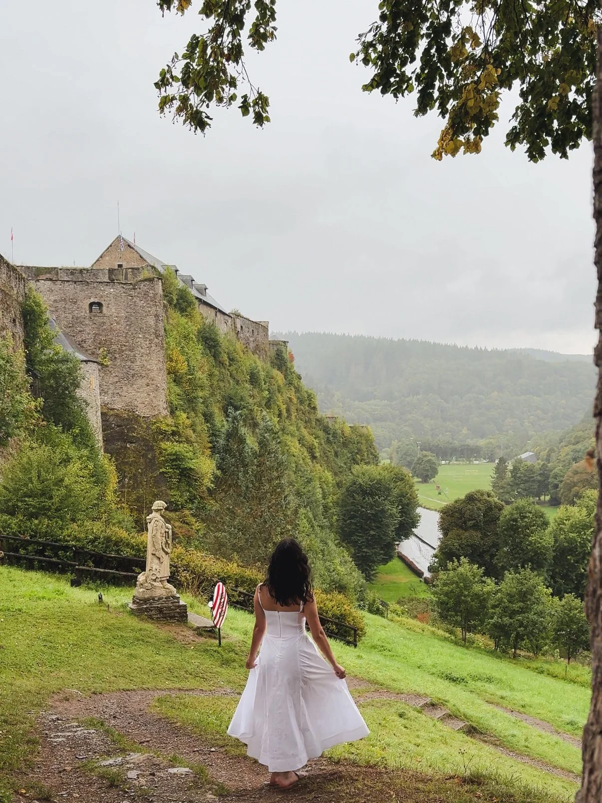A woman in a white dress standing outdoors on a grassy area, facing away from the camera, with a historic stone castle on a hill and a river in the background.