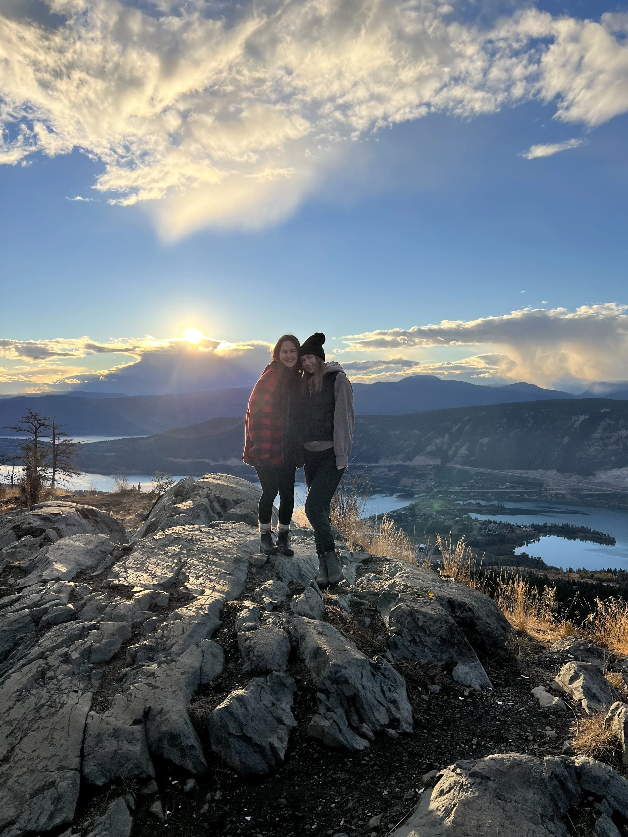 Two women in warm clothing smiling and standing on rocky terrain with a landscape of mountains, water, and a sunset sky in the background.