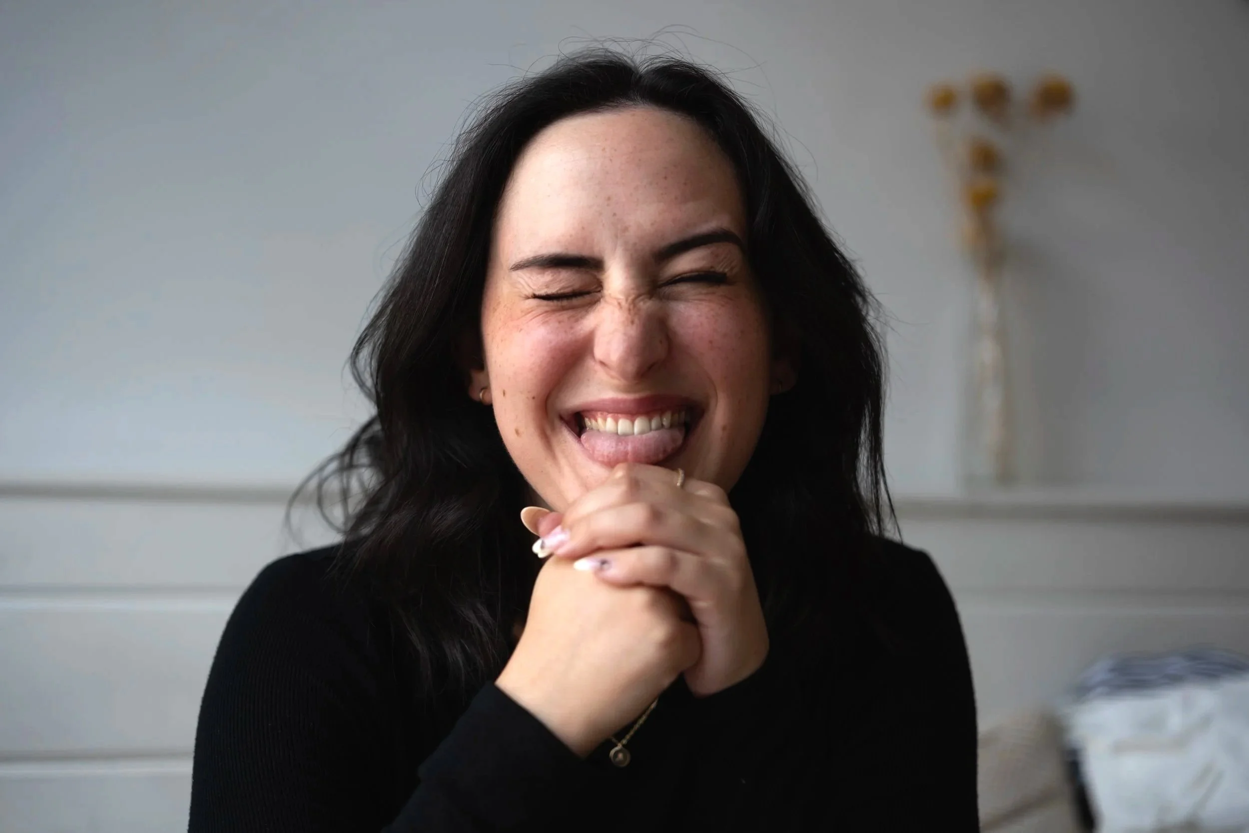 A woman with dark hair and freckles laughing with her eyes closed, hands clasped near her chin, indoor setting with gray wall and blurred gold-colored decoration in the background.