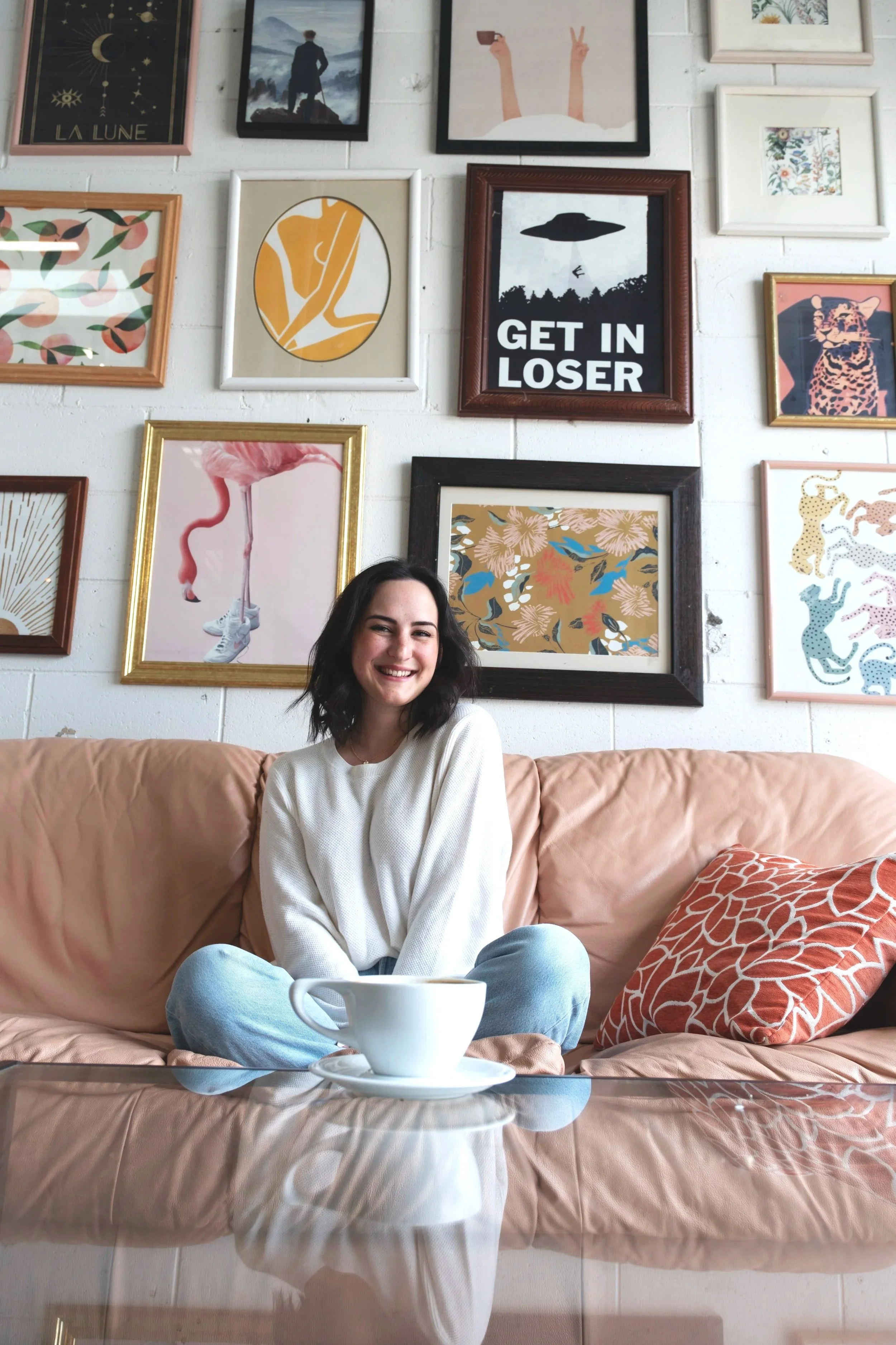 A young woman with dark, shoulder-length hair, sitting on a peach-colored sofa in a cozy, decoratively styled room with a coffee cup in front of her. The wall behind her features a gallery of framed artwork, including abstract, artistic, and humorous prints, with one prominently displaying the words "GET IN LOSER" alongside a silhouette of an UFO and trees.