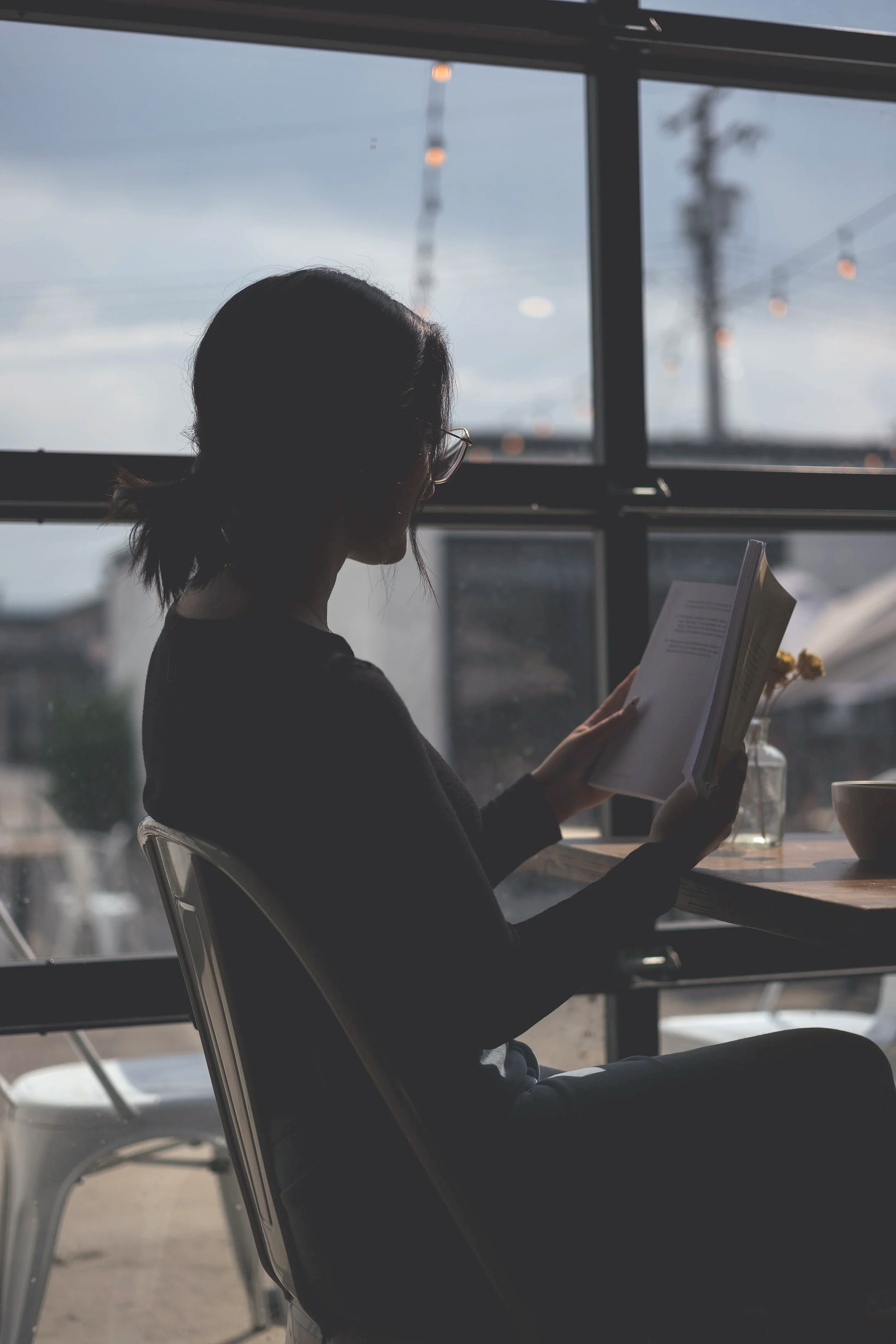 A woman with dark hair tied back, wearing glasses, sitting indoors near a large window, reading a book. The window shows a cloudy sky and a coffee shop outside.