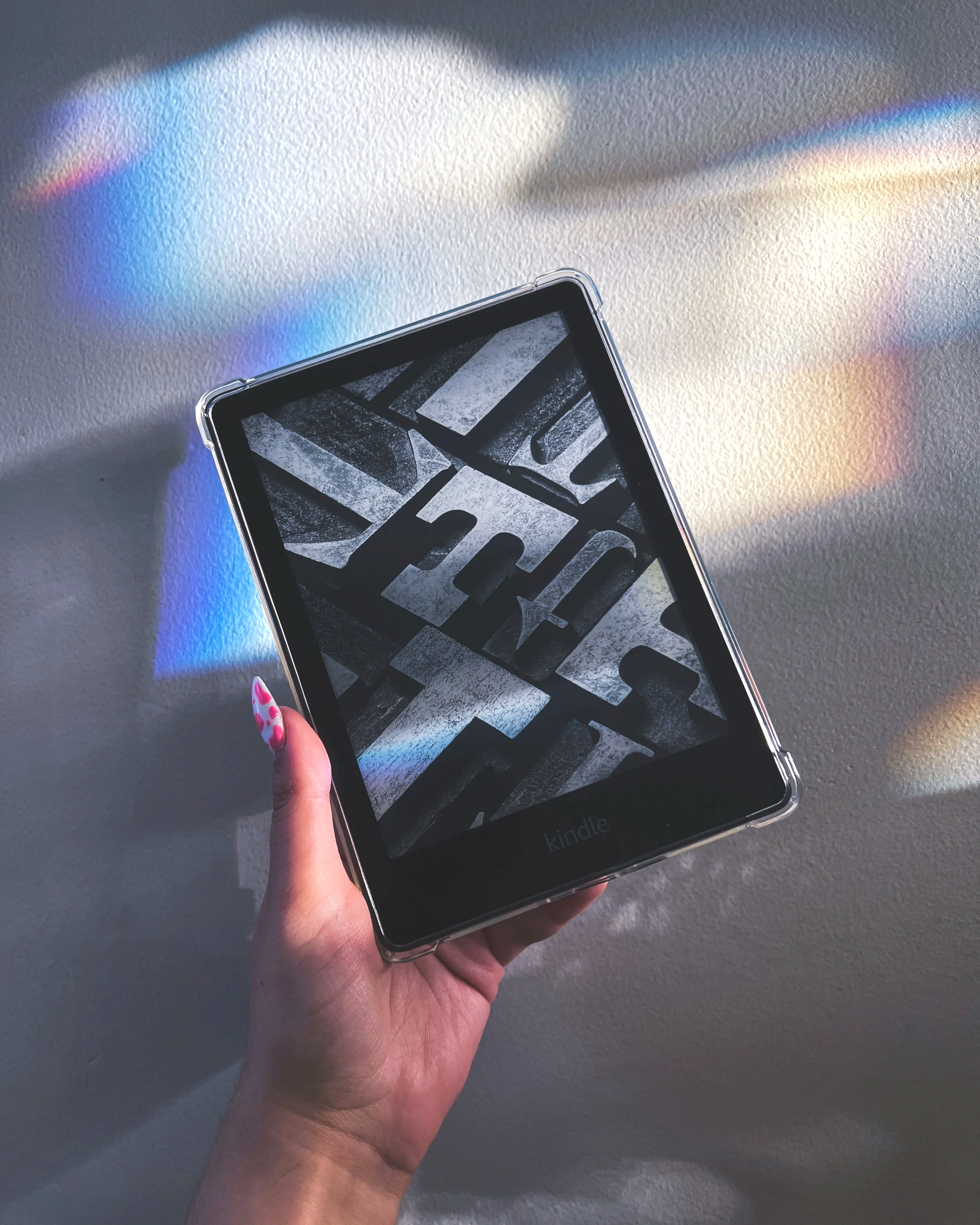 Close-up of a person holding a Kindle e-reader with a black-and-white geometric pattern on the screen, against a white wall with rainbow-colored light reflections.