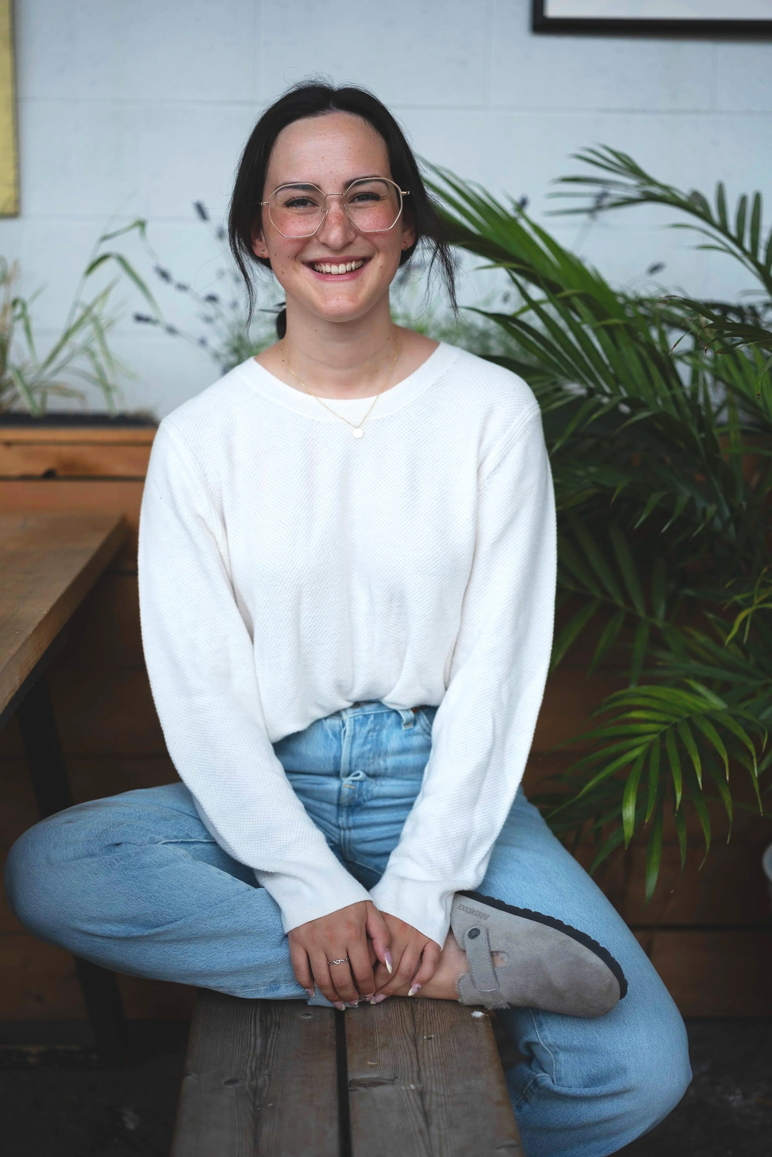 A smiling woman with dark hair and glasses, wearing a white sweater and blue jeans, sitting cross-legged on a wooden bench with green plants in the background.