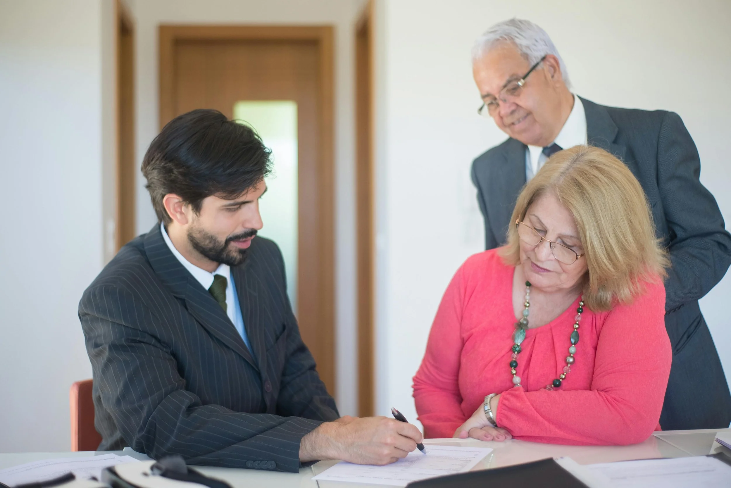 A business meeting with three people, a man and two women, gathered around a table, reviewing documents, with one woman wearing a pink top, another woman with glasses, and a man in a suit with a beard.