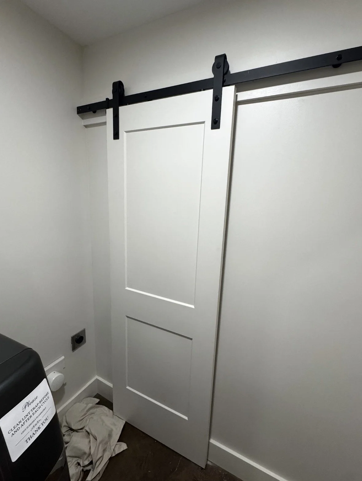 Sliding barn door on a black track in a room with white walls and a laundry machine in the corner.