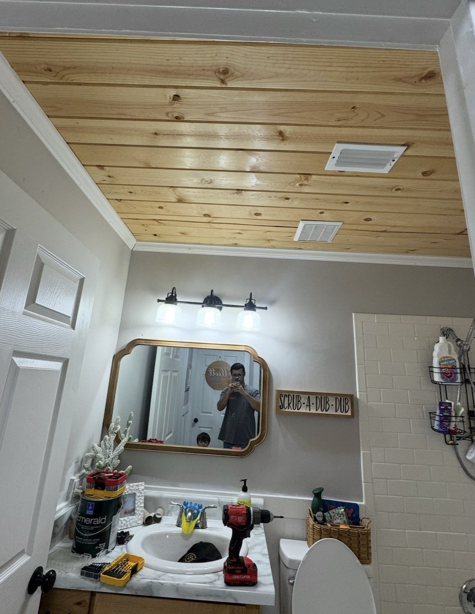 A bathroom with a wood-paneled ceiling, a mirror, a sink with tools and supplies on the counter, and a sign that reads 'Scrub-a-Dub-Dub.'