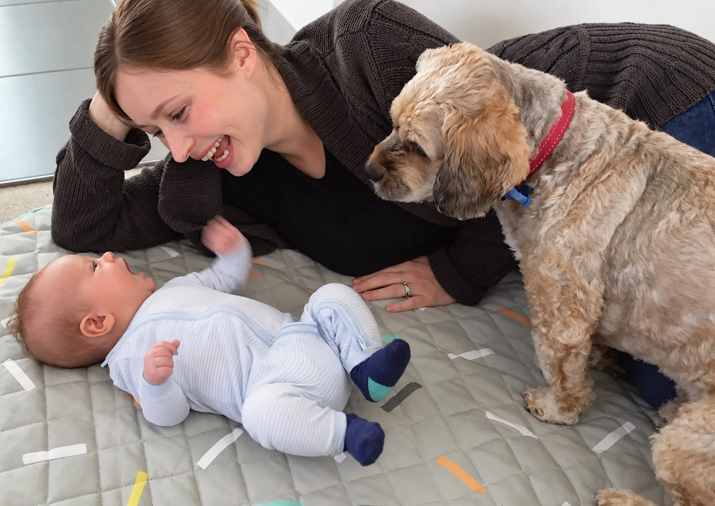 Smiling woman lying beside a baby and a curly-haired dog on a quilted play mat