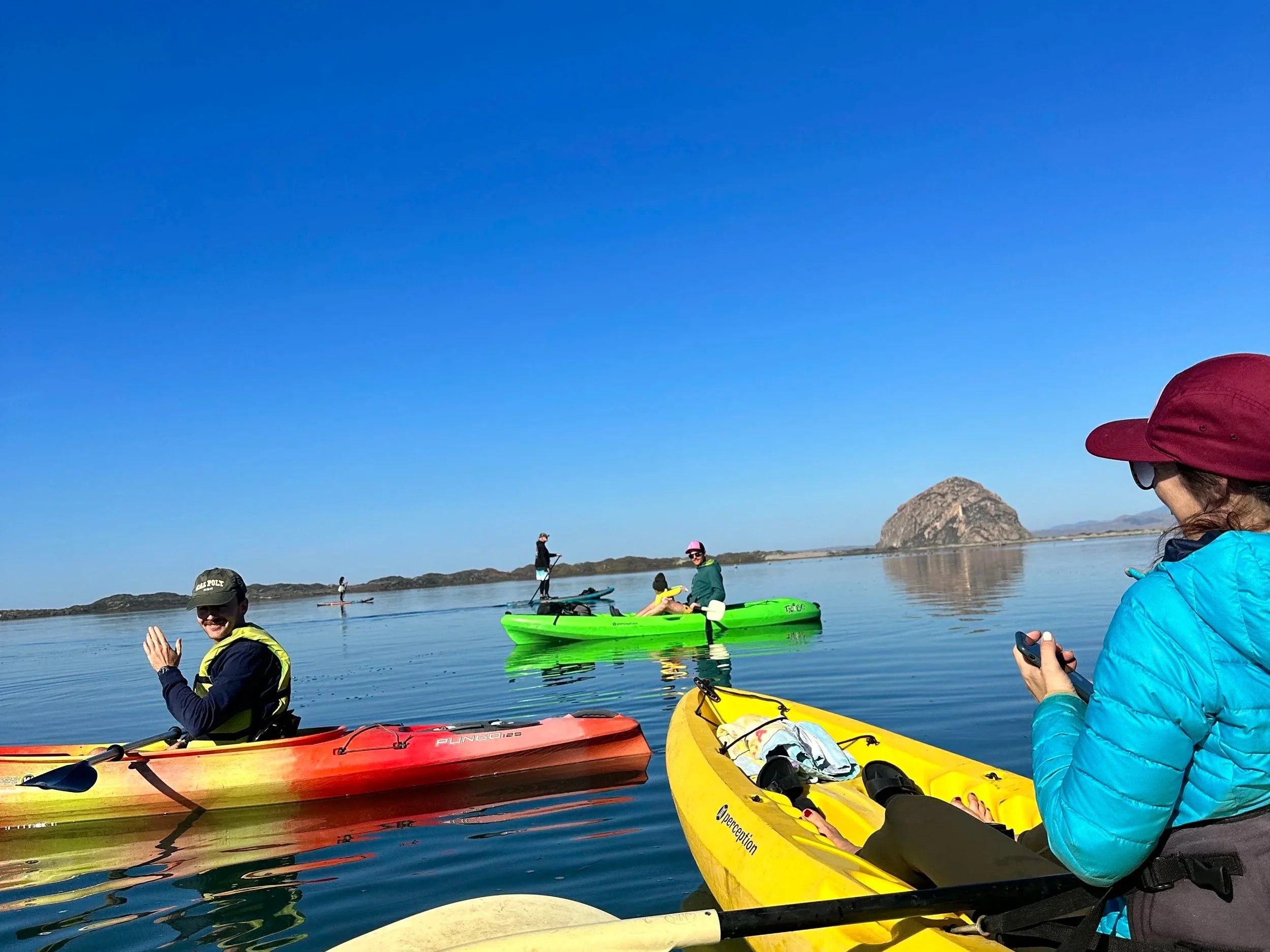 People kayaking on a calm lake with a rocky island in the background, clear blue sky, and individuals wearing life jackets and sun hats.