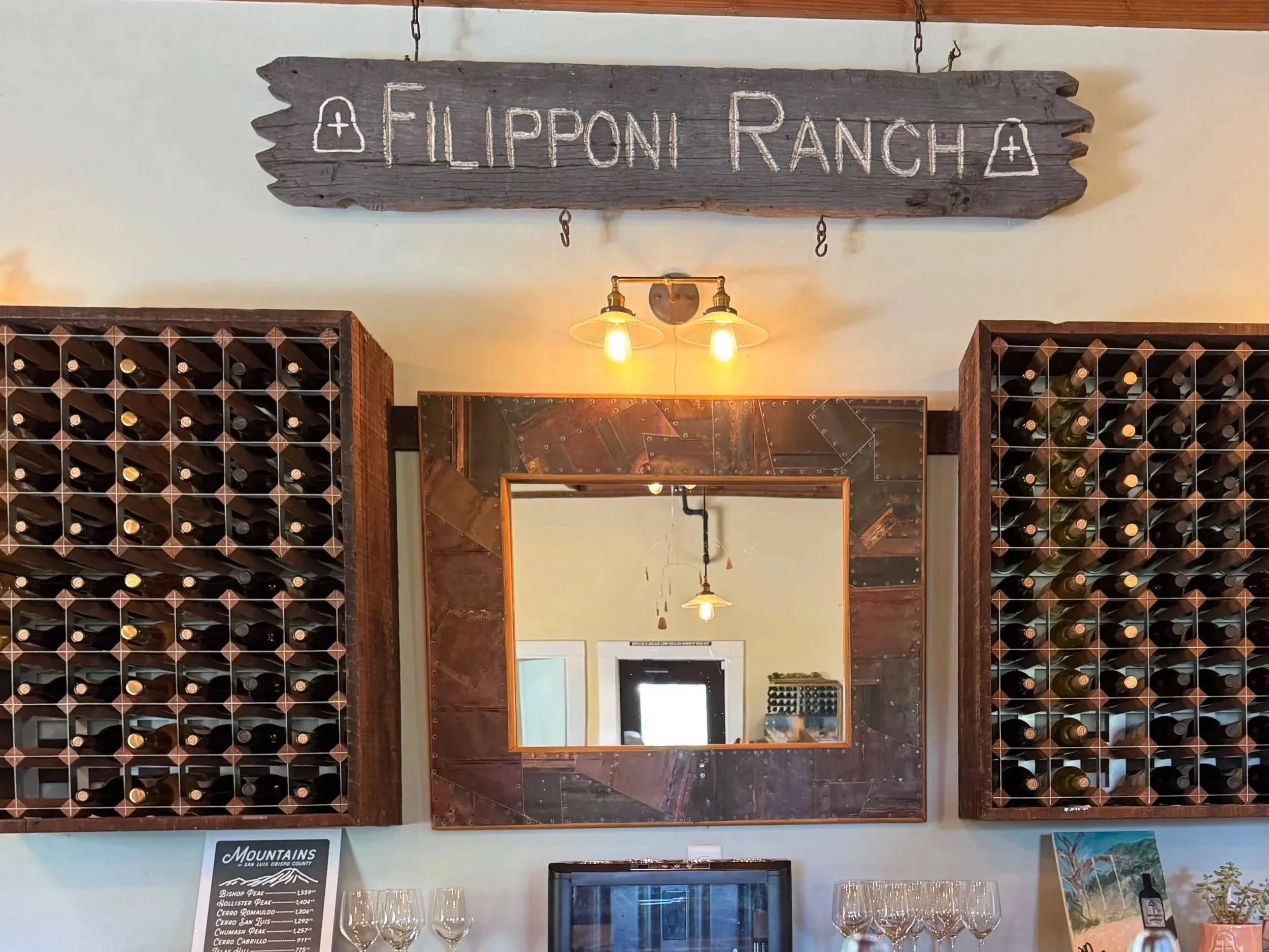 Wooden sign with white lettering and symbols reading 'Lippan Ranch' hangs above wine bottle racks, with warm yellow ceiling lights and a mirror below.