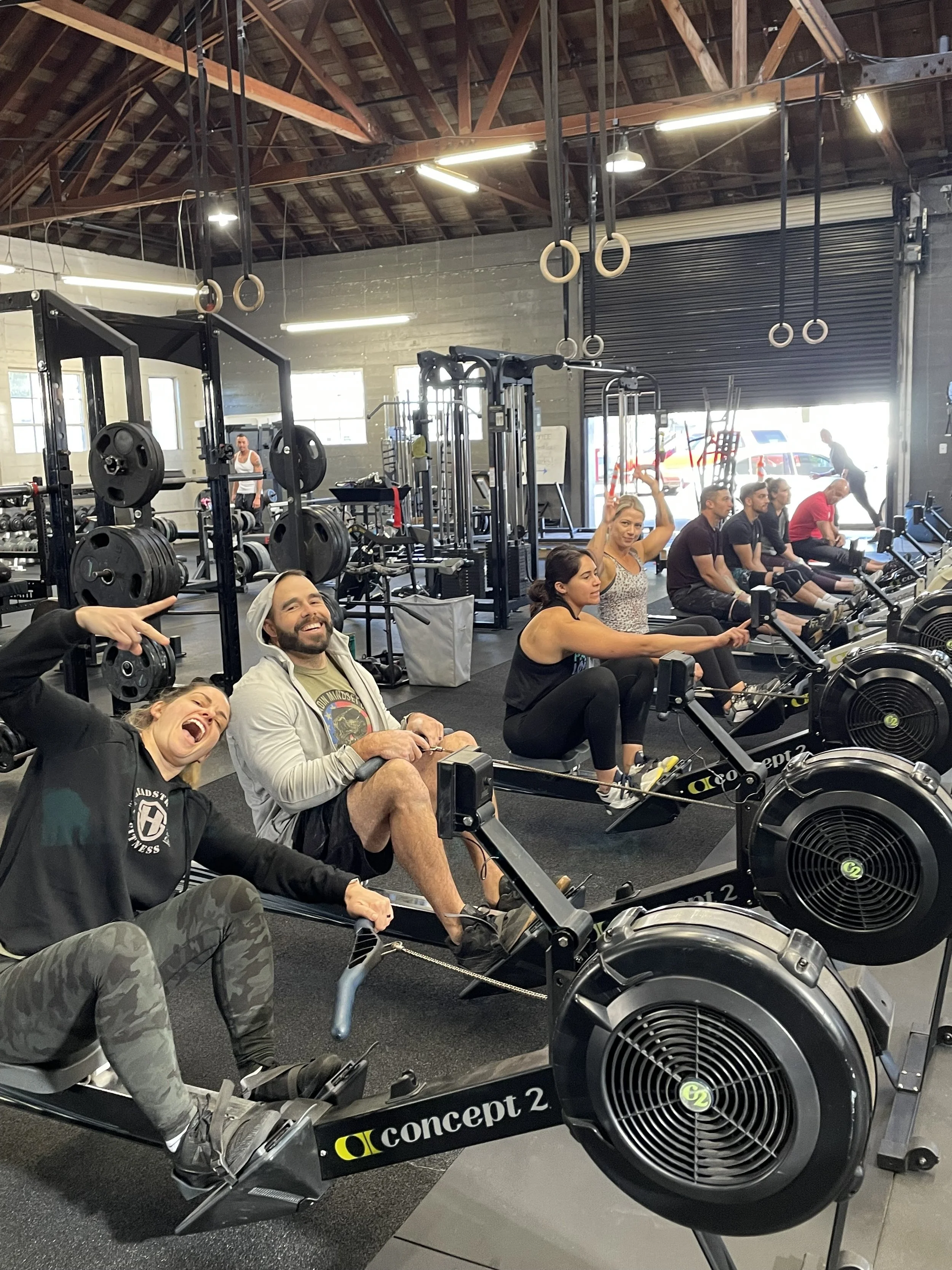 People working out on rowing machines inside a gym, with gym equipment and gymnastic rings hanging from the ceiling.