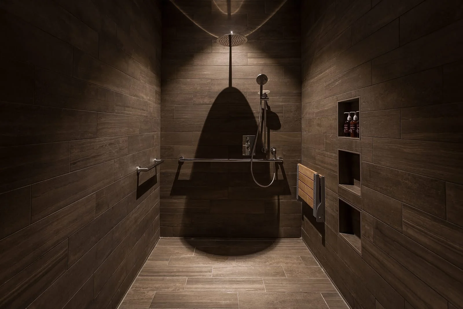 A modern shower with brown tiled walls and a wooden bench, featuring a rain showerhead and wall-mounted shower controls. There are built-in shelves holding bottles and a towel bar.