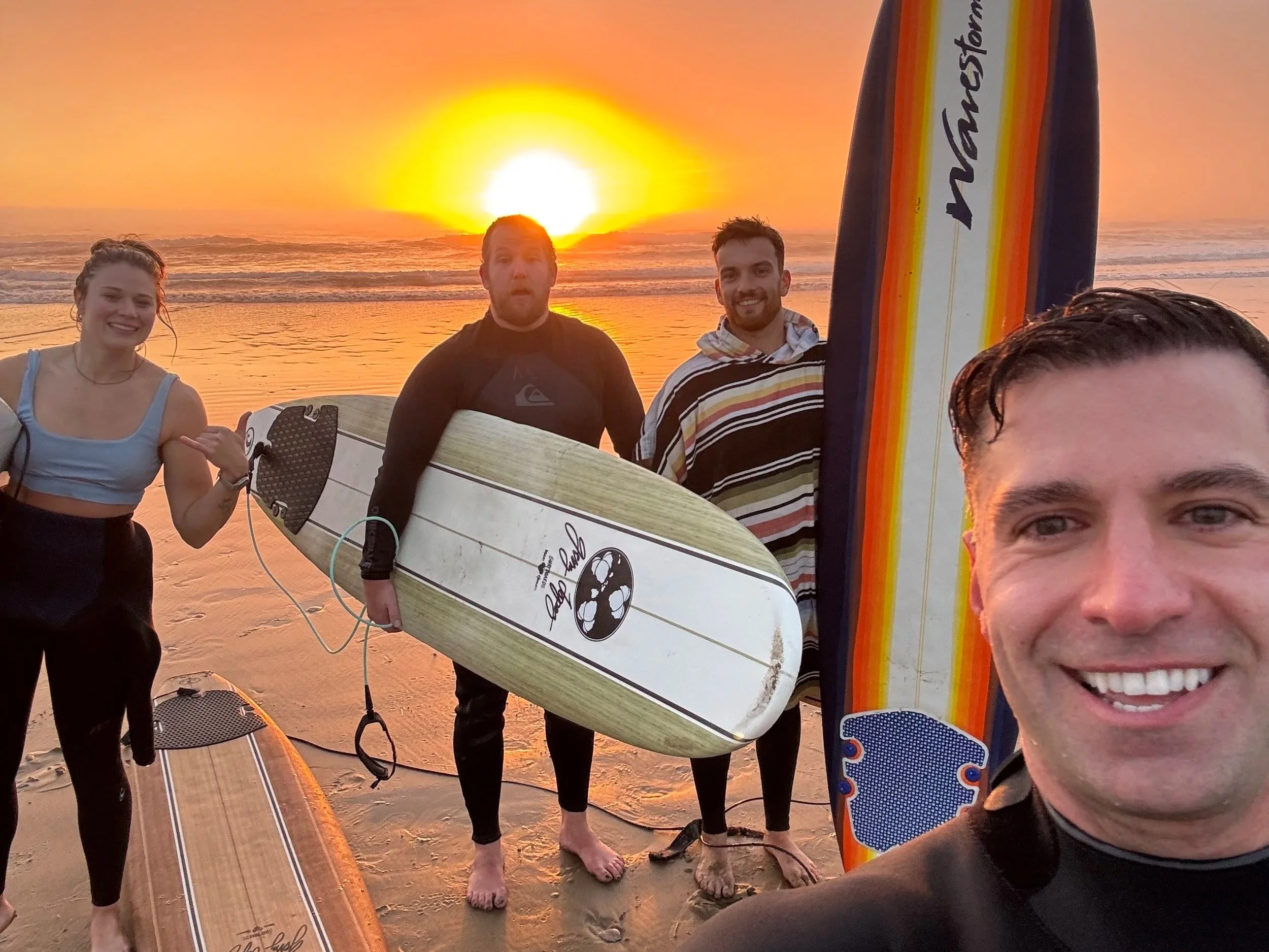 A group of people at the beach during sunset, holding surfboards and smiling.