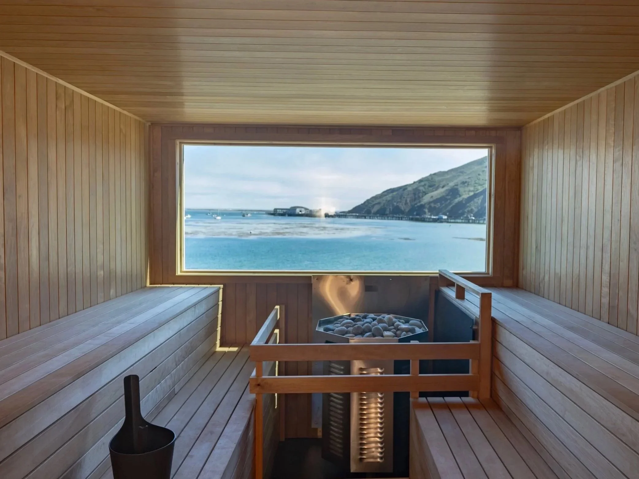 Interior of a wooden sauna with a large window showing a scenic ocean view with mountains, boats, and a pier.