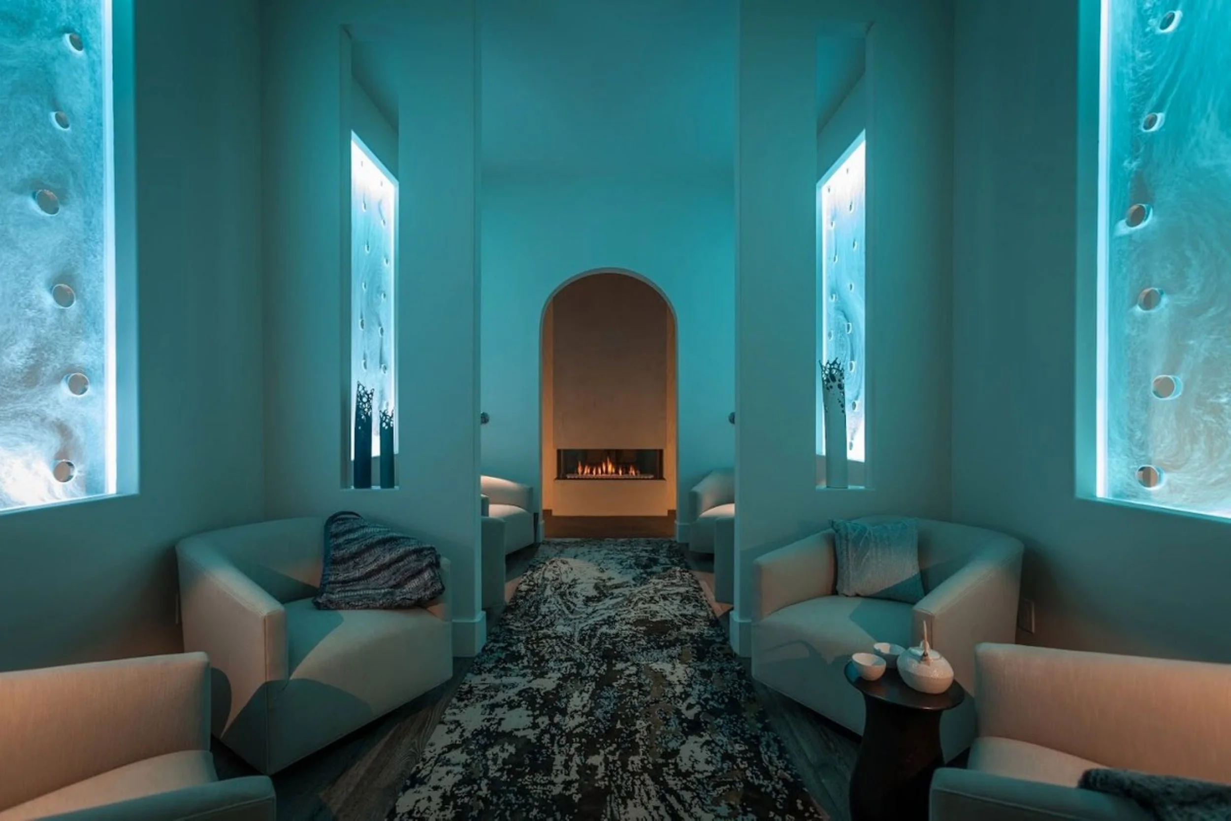 A modern living room with light-colored curved sofas arranged in a circular formation, a patterned rug, a fireplace with a fire inside, and tall, narrow windows with water-themed artwork, illuminated with blue lighting, on the walls.