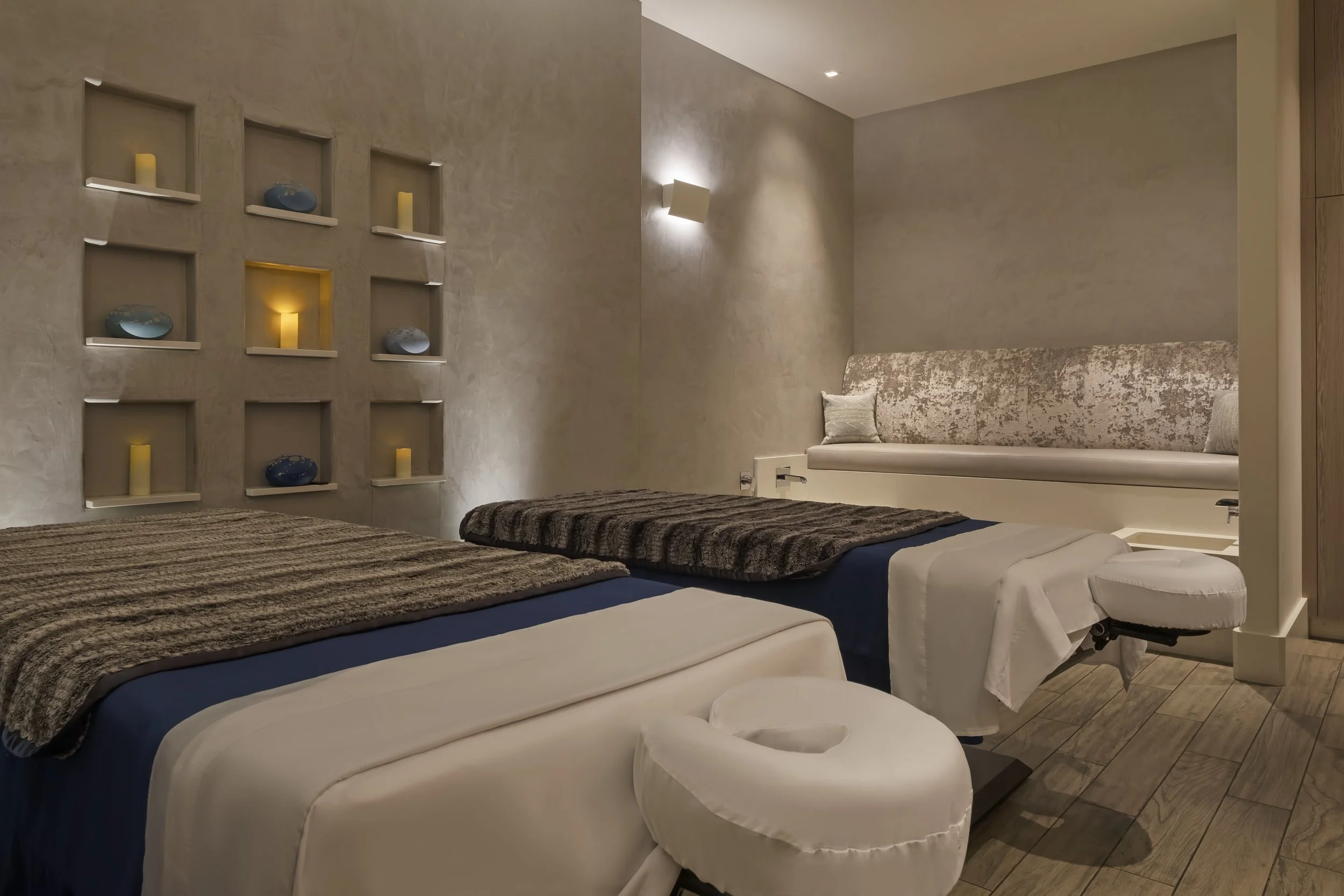 A hotel room with twin beds, white bedding, and dark brown throws. A beige bed bench is at the foot of the beds. The wall behind has built-in shelves with candles and decorative items. There is a beige textured wall with a wall-mounted light fixture. The floor is wood, and there is a cream-colored, textured accent wall on the right.