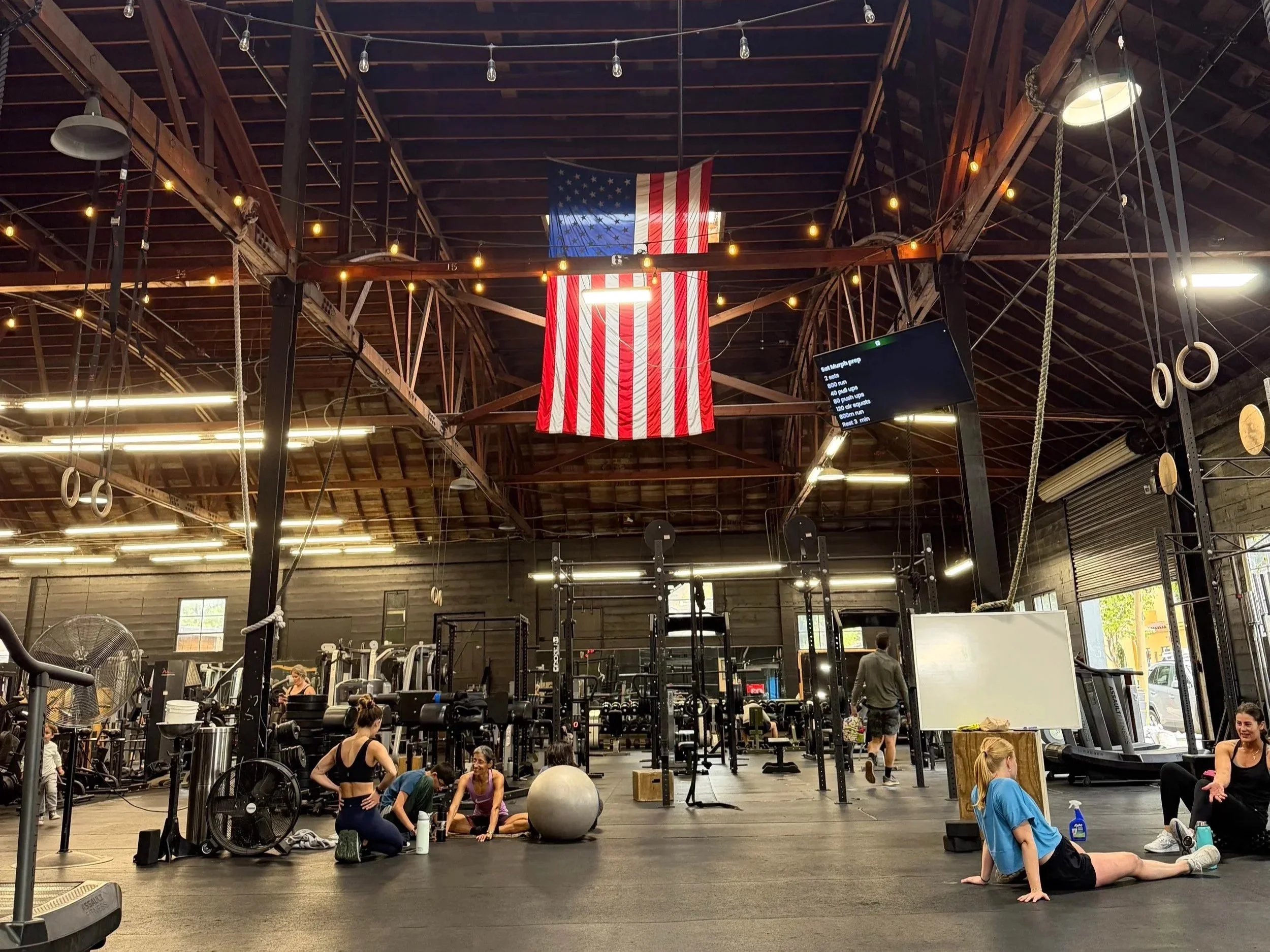 People exercising in a gym, with women stretching on the floor, weights and gym equipment around, an American flag hanging from the ceiling, and gymnastic rings hanging from above.